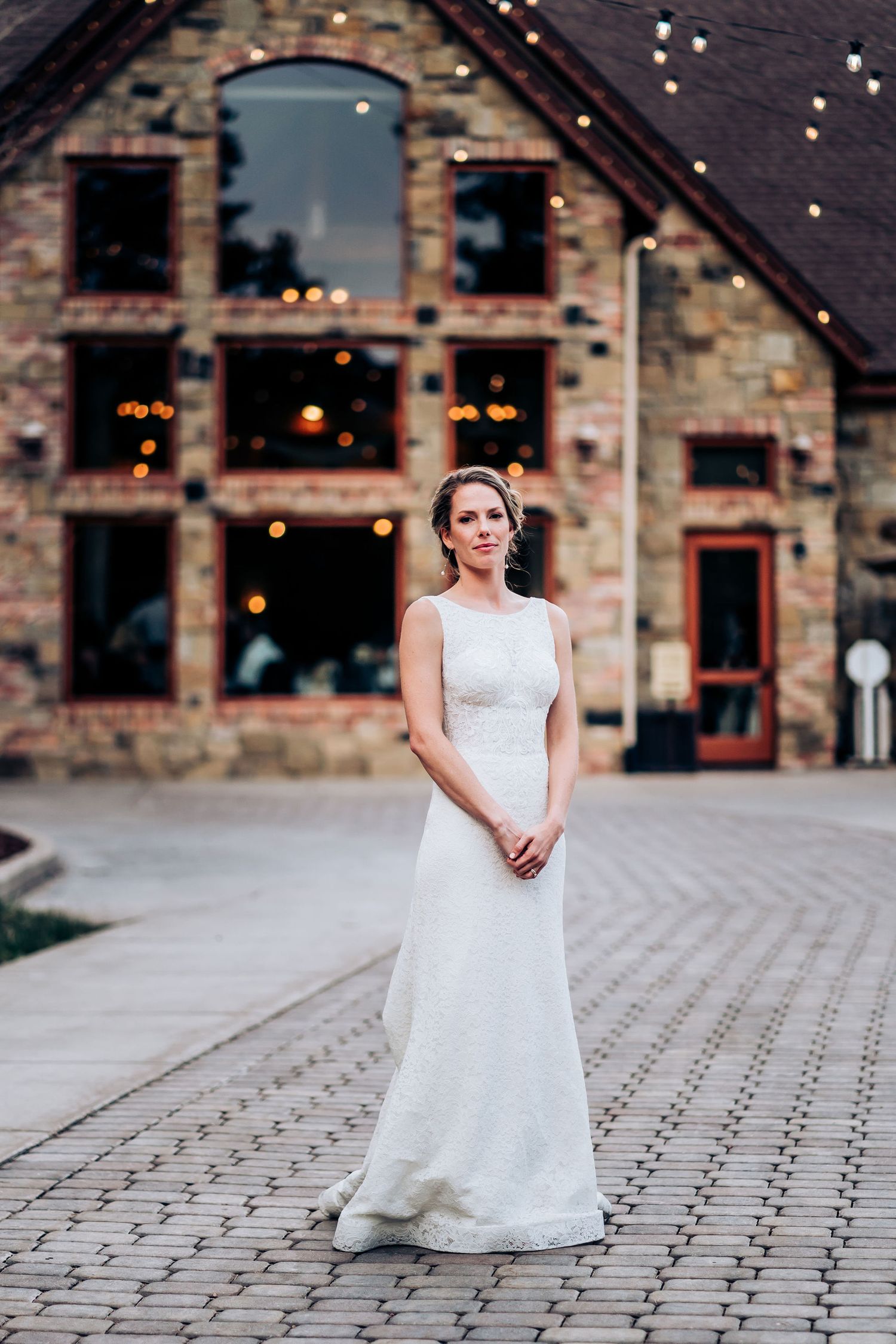 A person in a sleek white wedding dress poses in front of a stone building with twinkling lights.