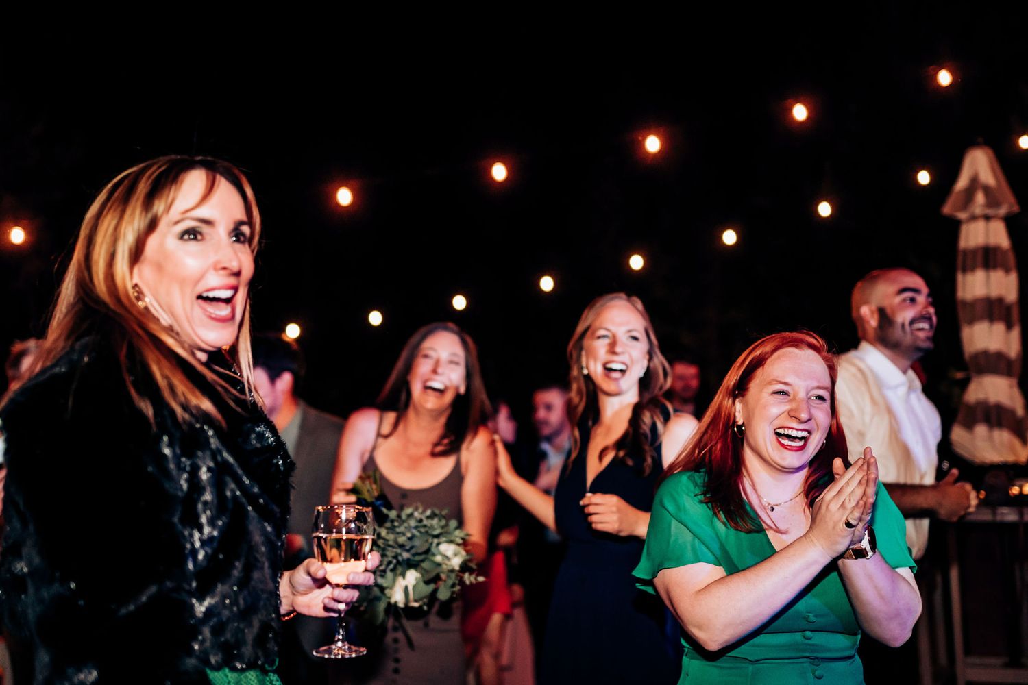 Joyful guests laughing and celebrating at nighttime wedding reception under string lights.