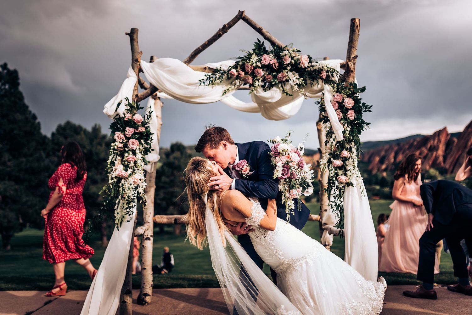 Romantic kiss under floral wedding arch with dramatic stormy sky and mountains.