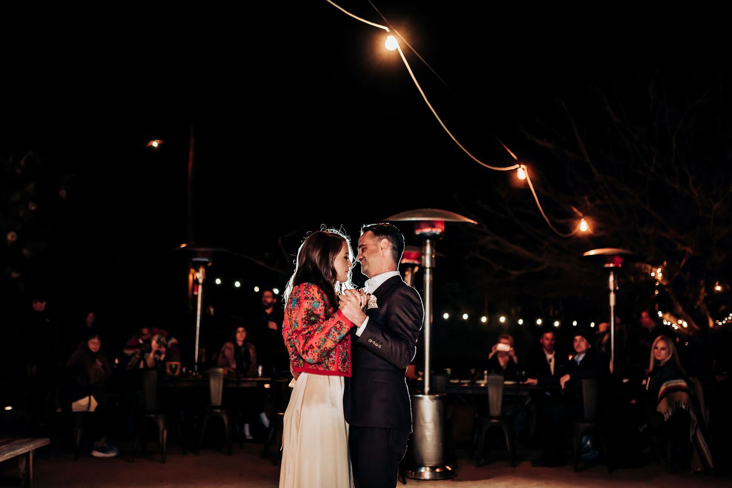 Couple dancing under string lights and heat lamps at an evening outdoor celebration.