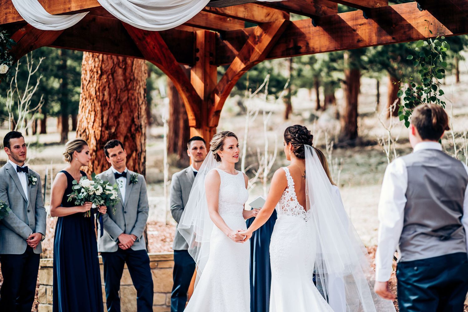 Outdoor wedding ceremony taking place under a wooden pergola with tall pine trees.