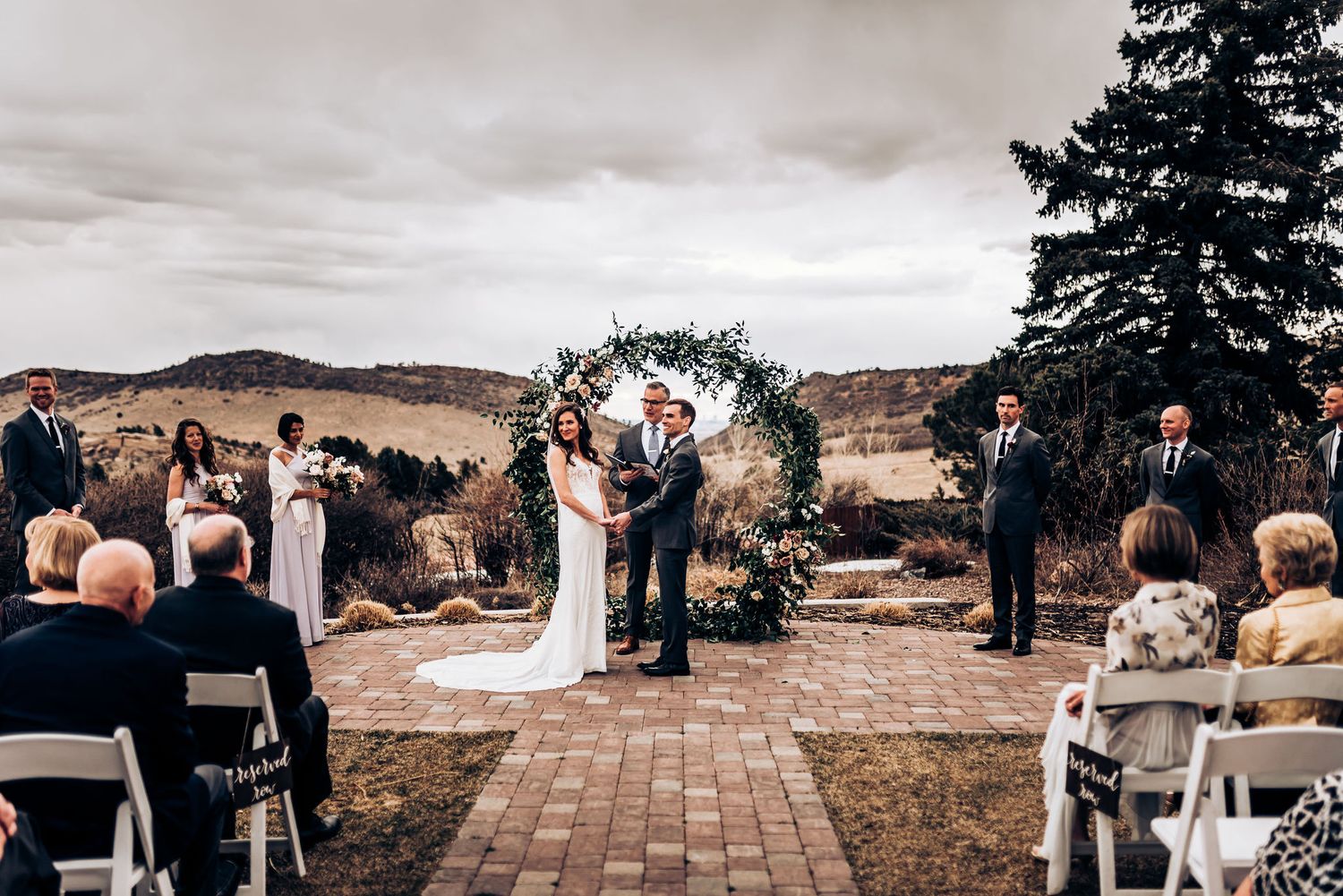 Outdoor wedding ceremony with guests seated on white chairs facing a decorated floral arch.