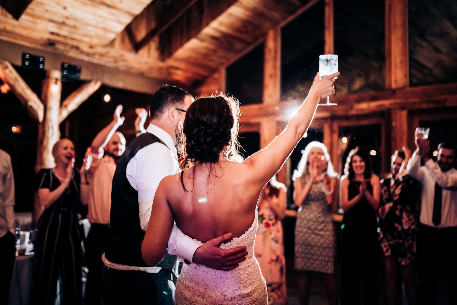 Couple dances while guests raise their glasses in celebration at rustic reception venue.