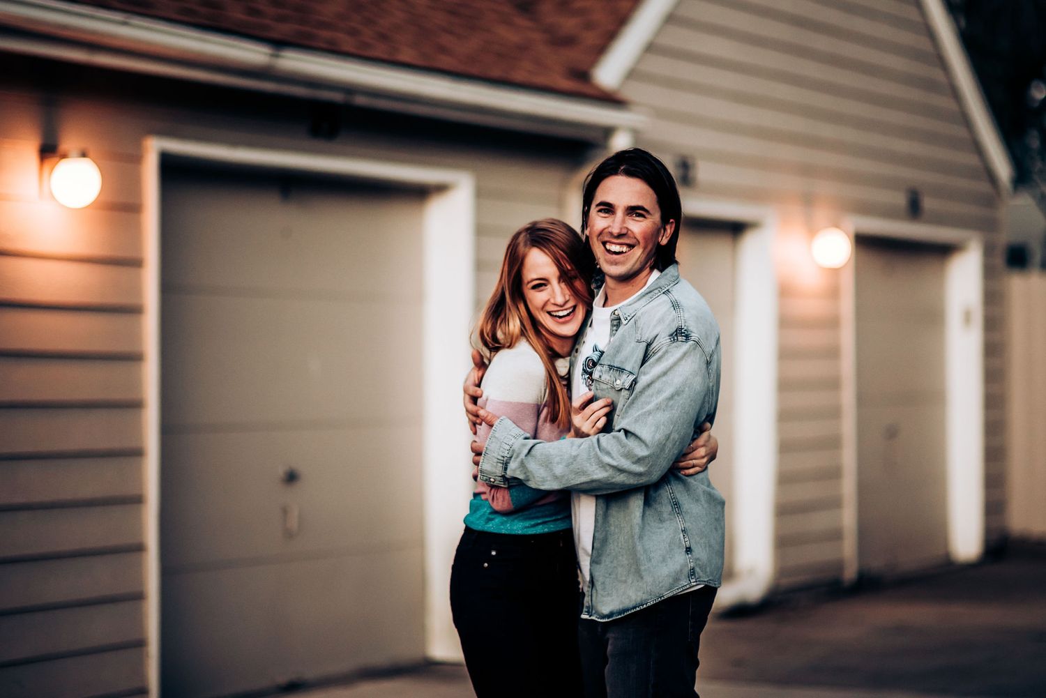 A romantic couple embraces and smiles outside garage doors at dusk with warm lighting.