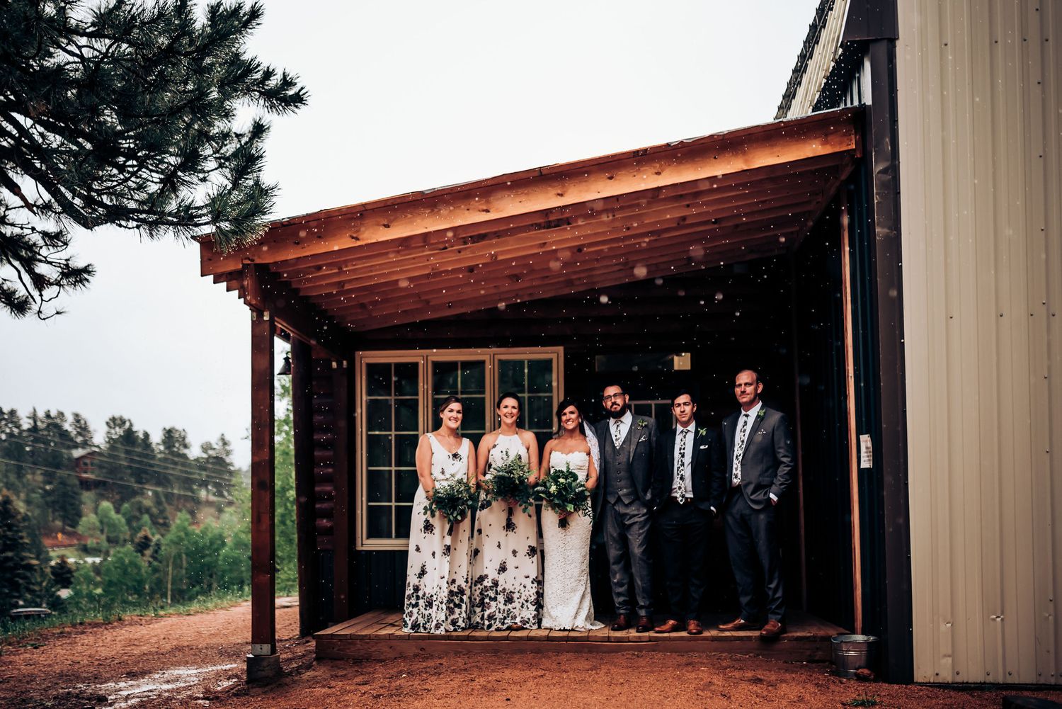 Wedding party stands under wooden overhang against mountainous backdrop.