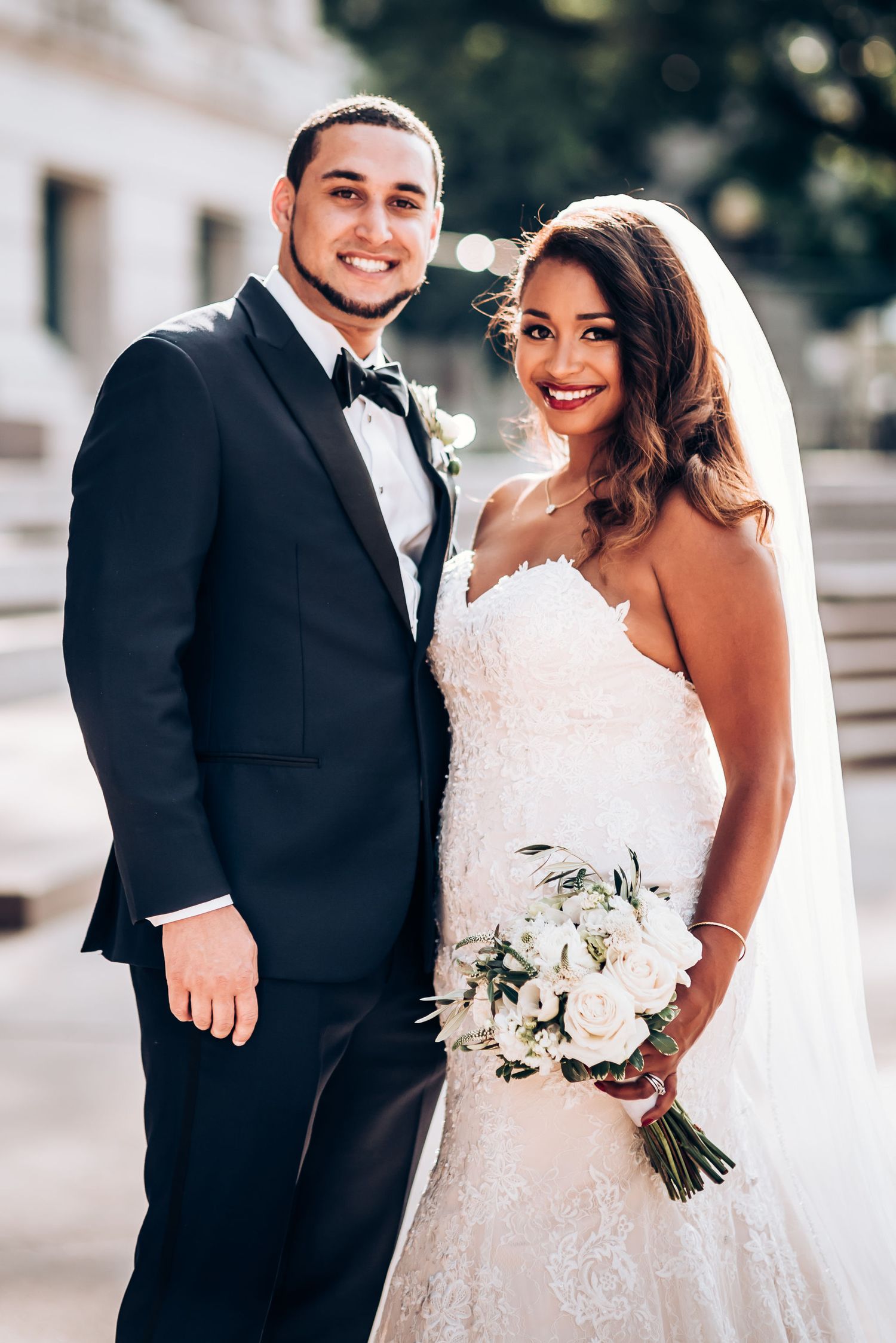 A wedding couple smiles for a portrait, with the bride holding a white flower bouquet and wearing a lace gown.