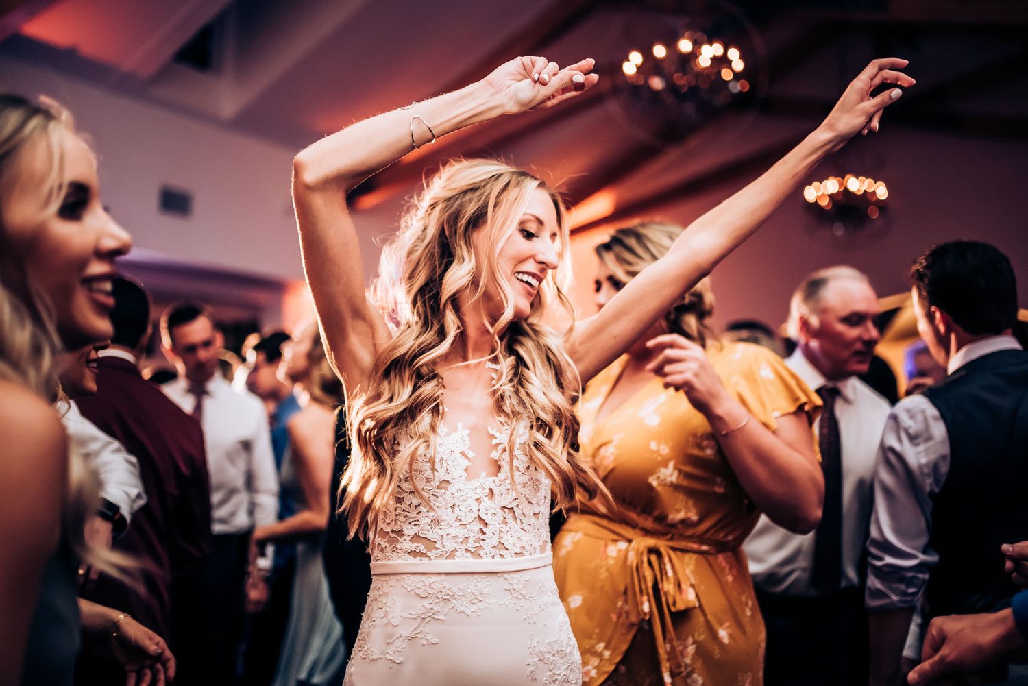A bride in a stunning white lace dress dances with raised arms during her wedding reception.