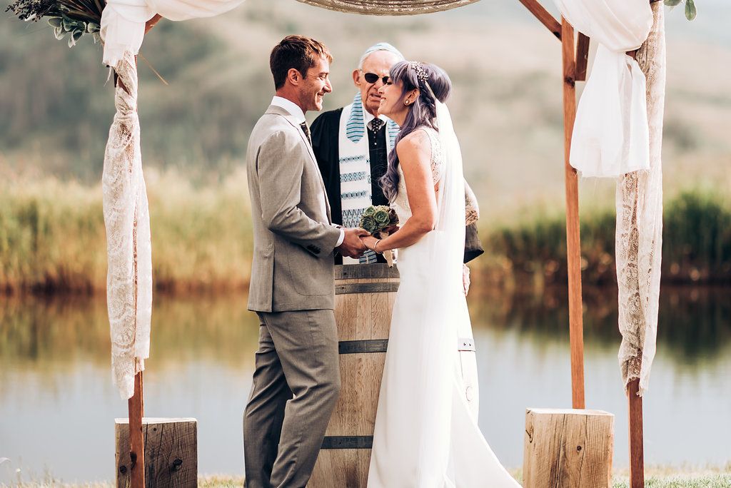 An intimate wedding ceremony by a lake with wooden posts and white fabric creating an elegant archway.