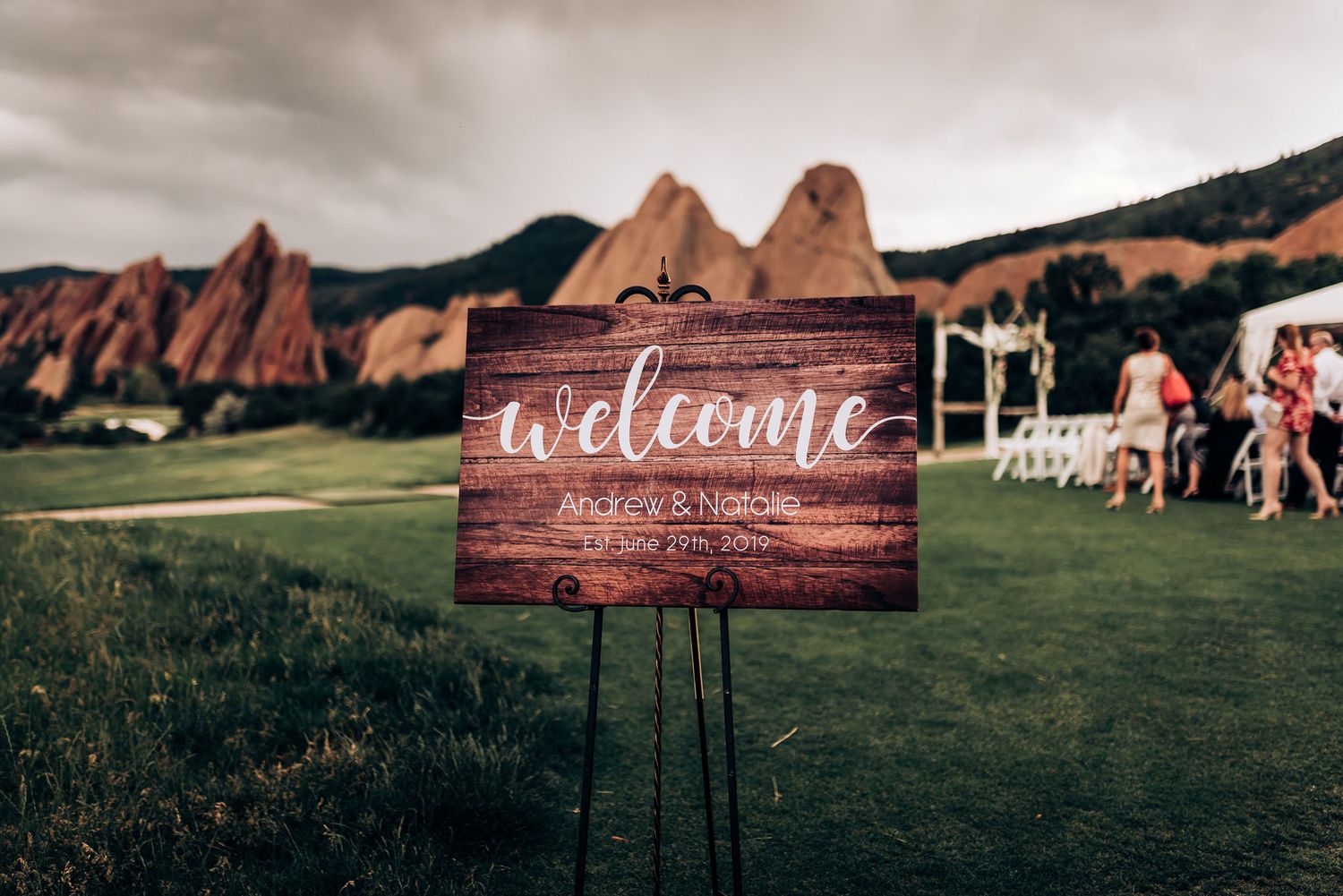Rustic wooden welcome sign stands in front of iconic red rock formations at outdoor venue.