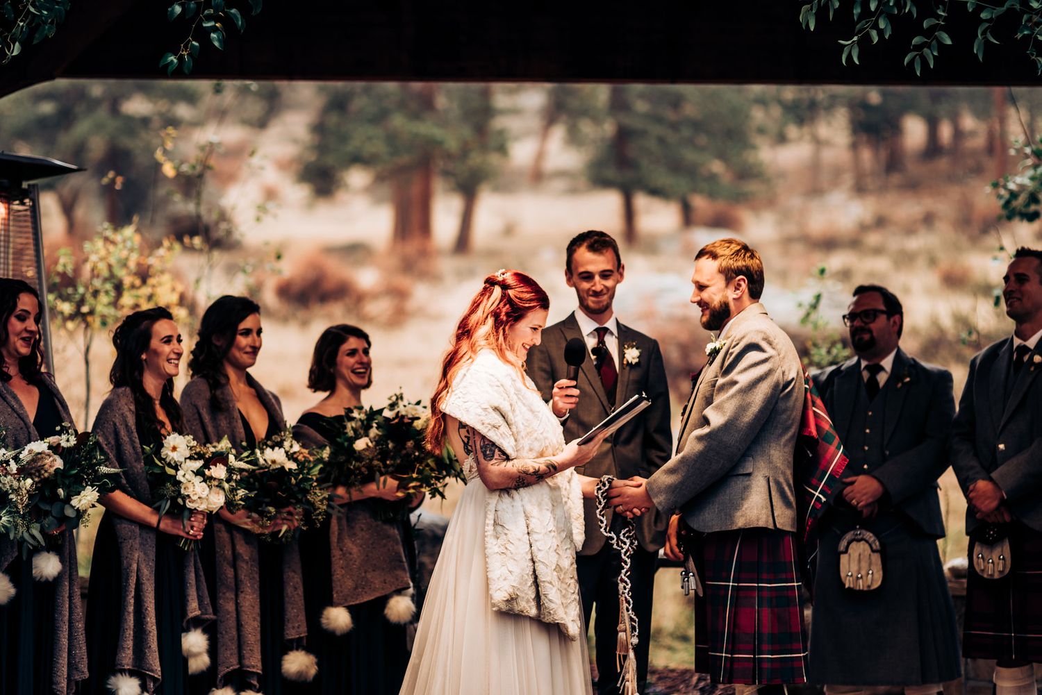Intimate outdoor wedding ceremony features couple exchanging vows surrounded by wedding party in kilts and gray dresses.