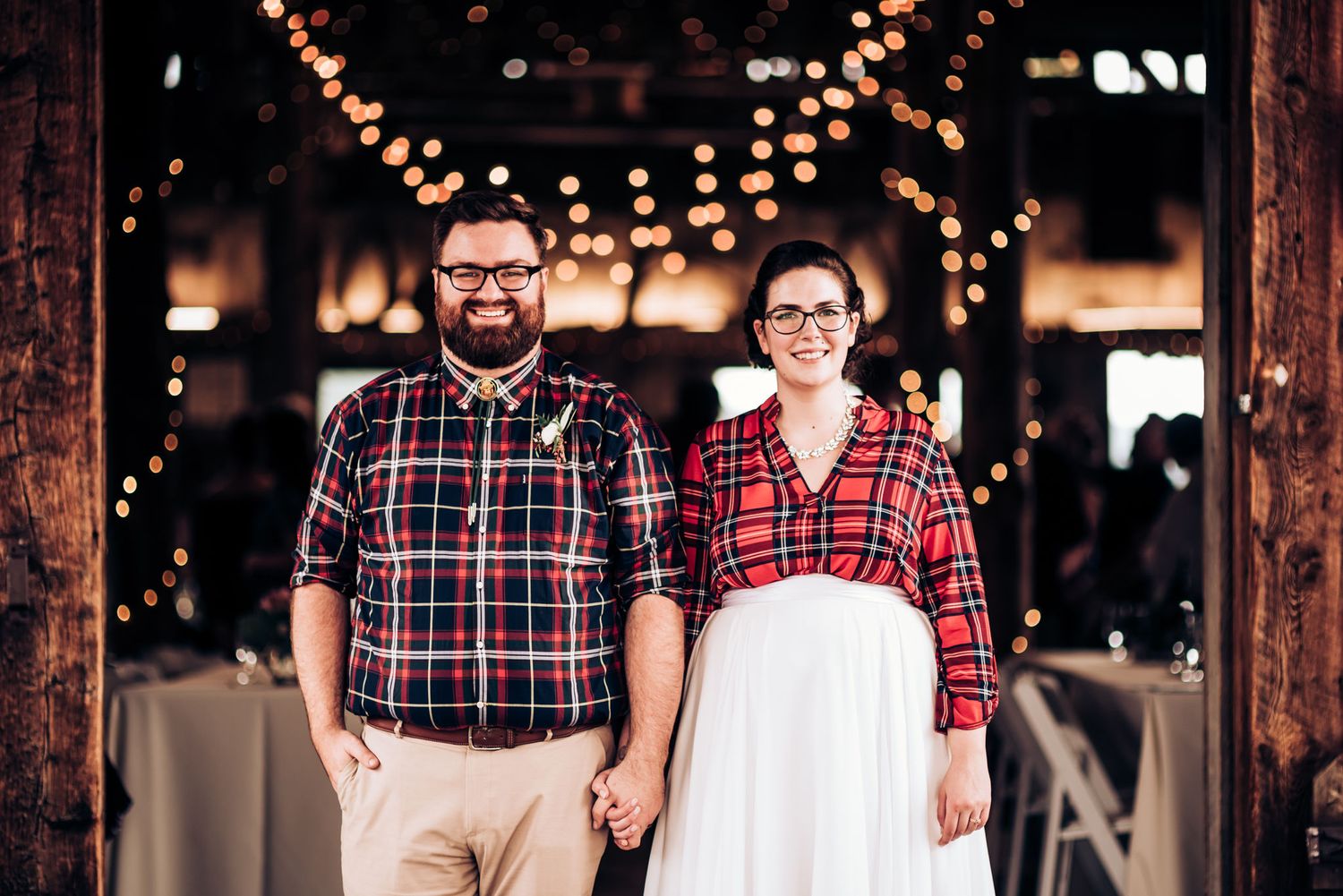 Couple in matching red plaid flannel shirts stand beneath string lights at indoor venue.