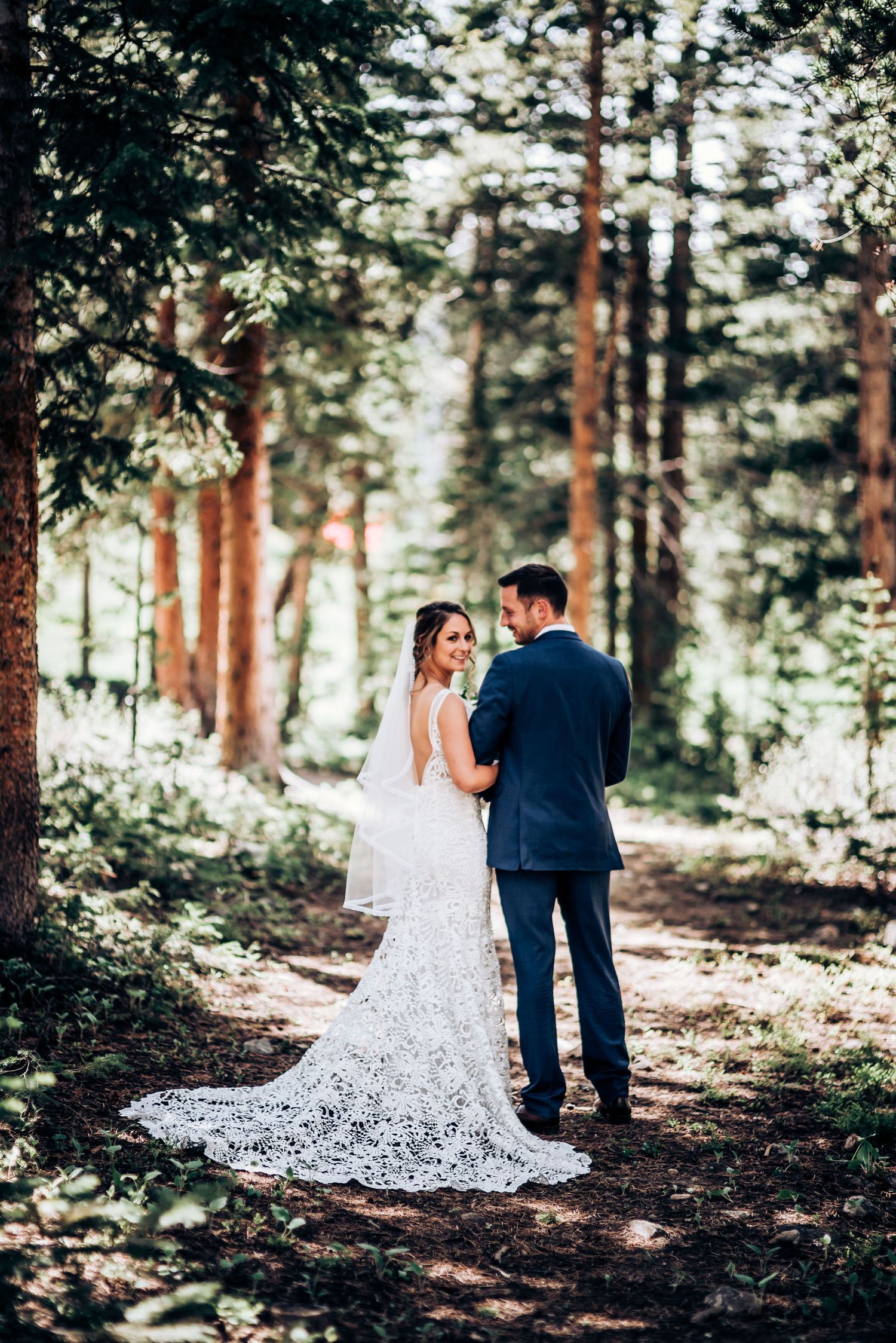 A stunning lace wedding dress train trails behind the couple on a forest path.