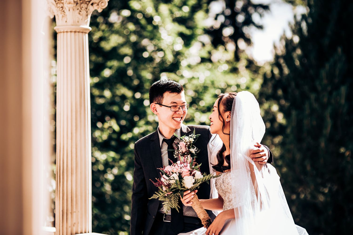 A romantic wedding portrait featuring a couple with floral bouquet under an ornate architectural column.