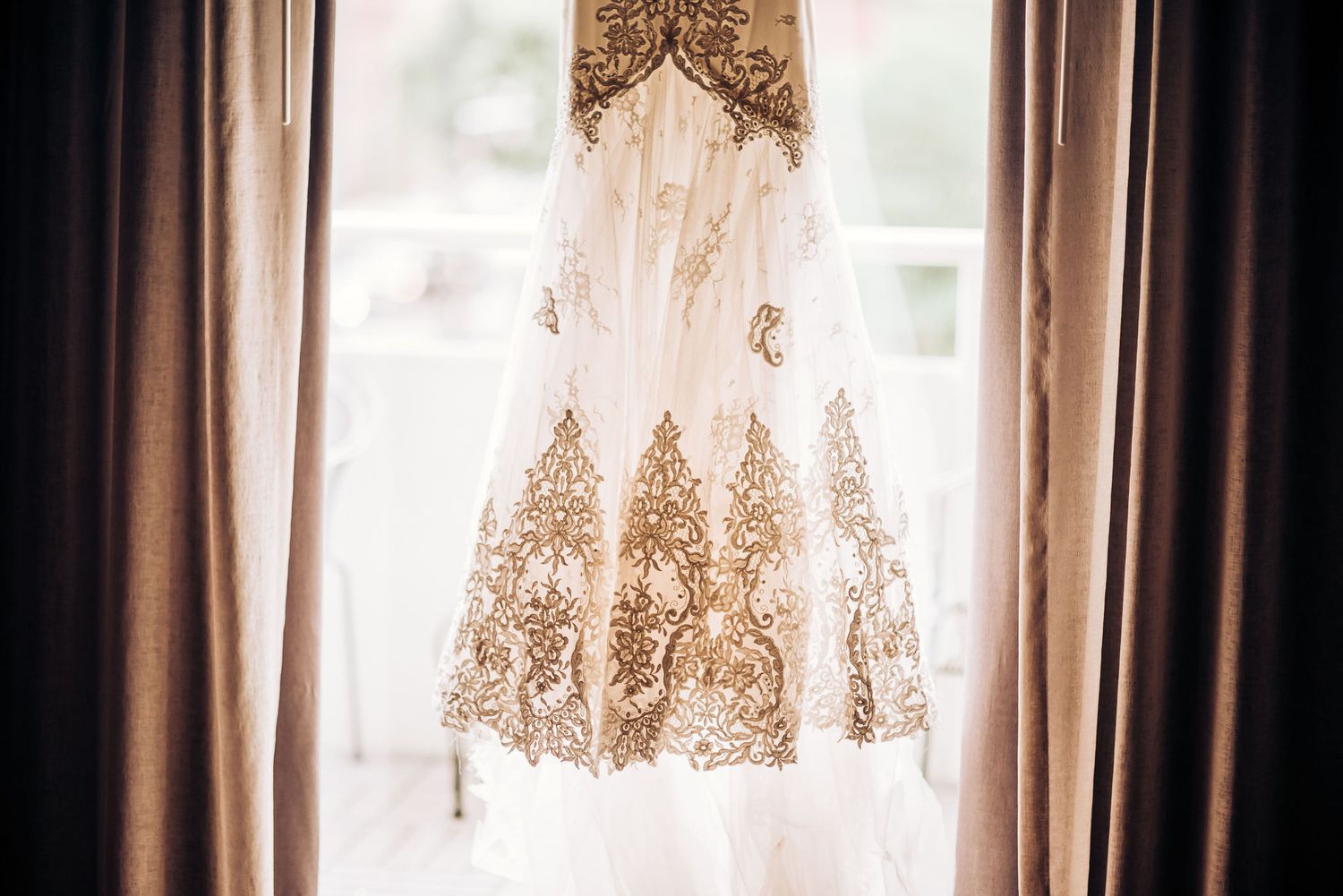 Ornate white wedding dress with lace details hanging in window light between curtains.