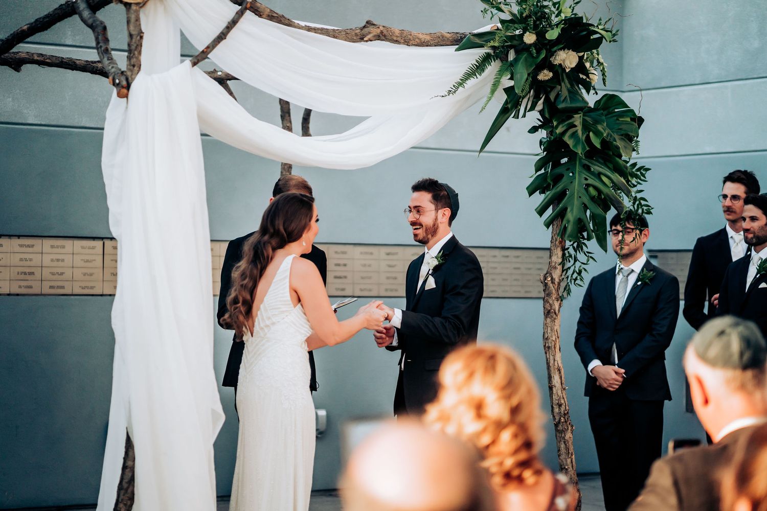 Wedding ceremony in modern venue with white draped arch and greenery decorations.