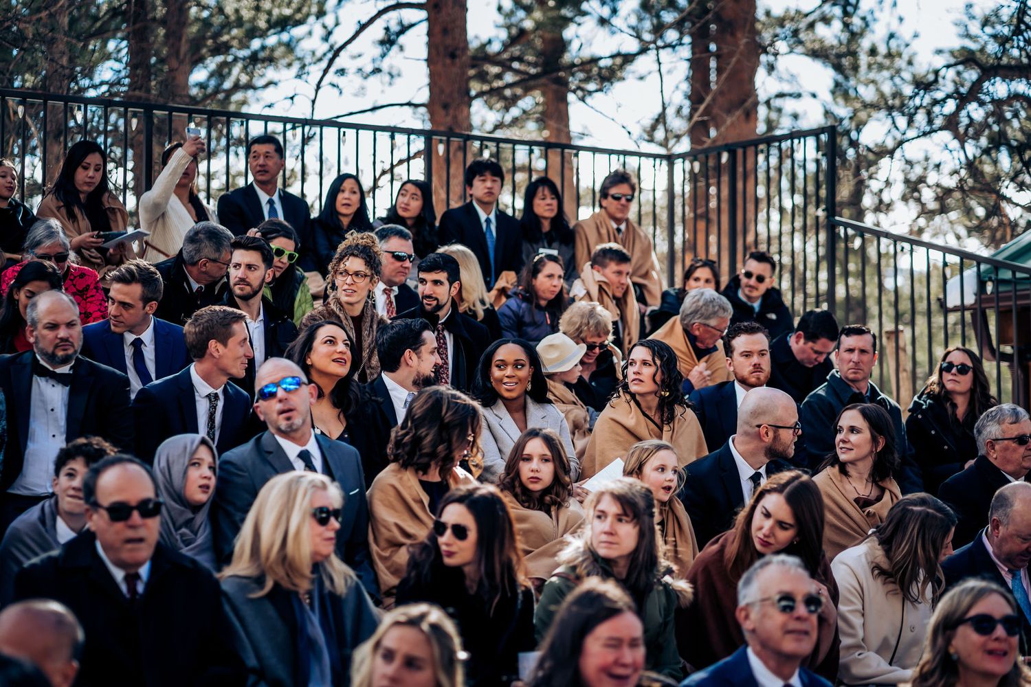 Well-dressed wedding guests seated in rows at an outdoor mountain venue with pine trees in background.