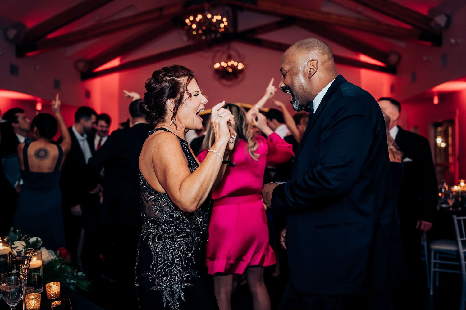 Guests dancing and singing enthusiastically at a wedding reception with red ambient lighting.