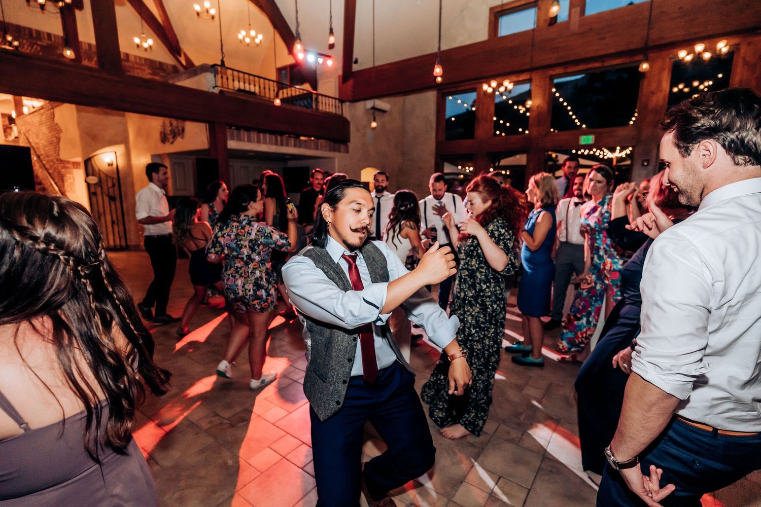 Wedding guests crowd the dance floor during a lively reception in a rustic barn venue.