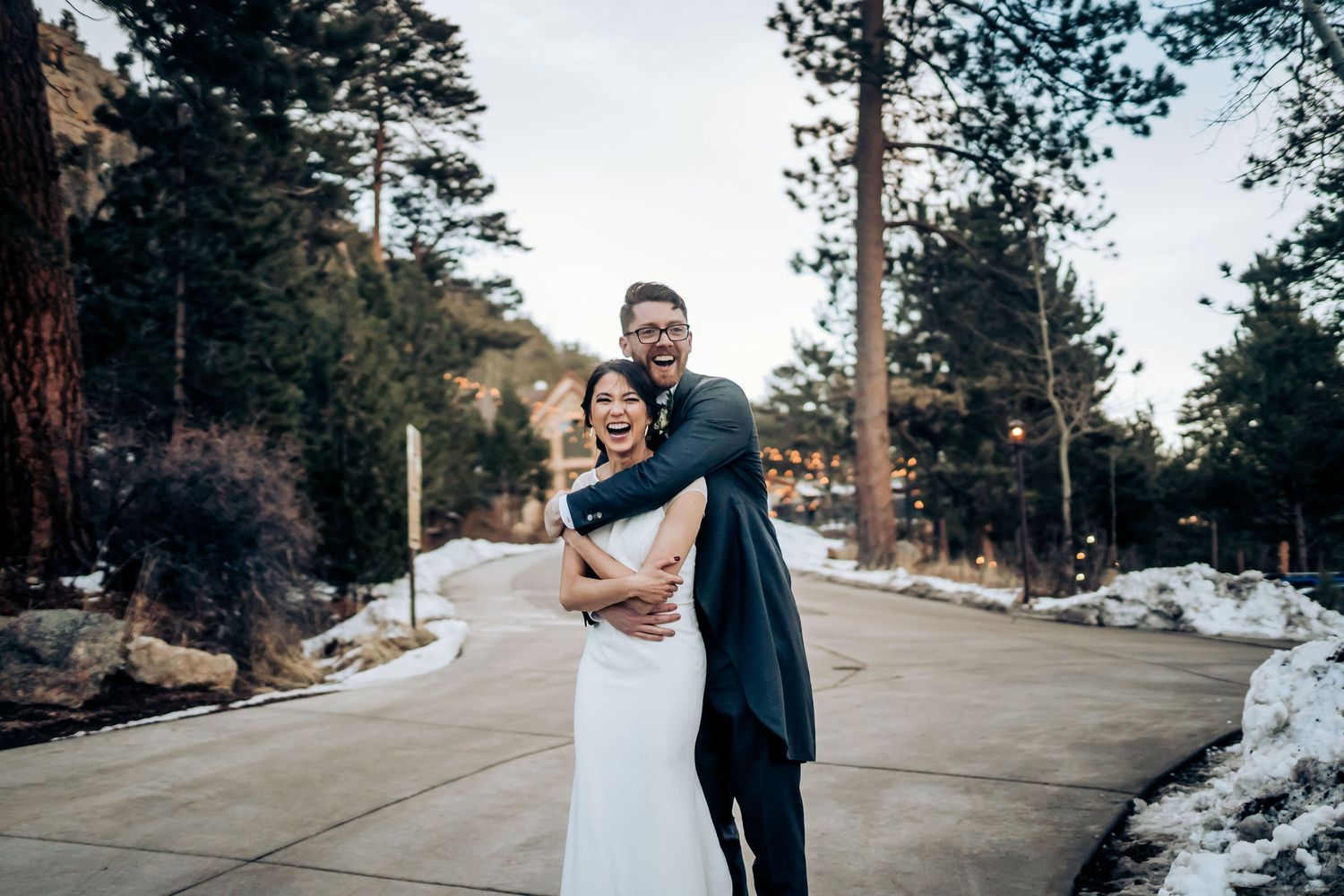 Couple embraces on a snowy pathway surrounded by tall pine trees.