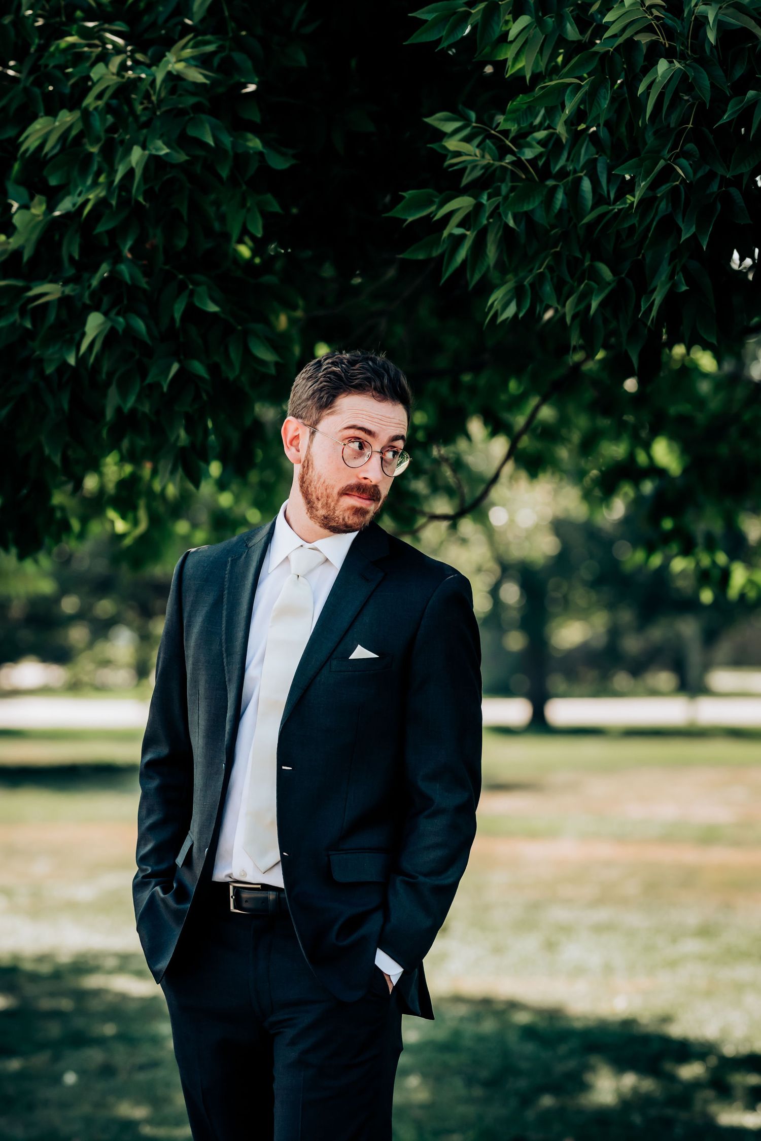 A formal portrait of a groom in a classic black suit and bow tie standing under green foliage.