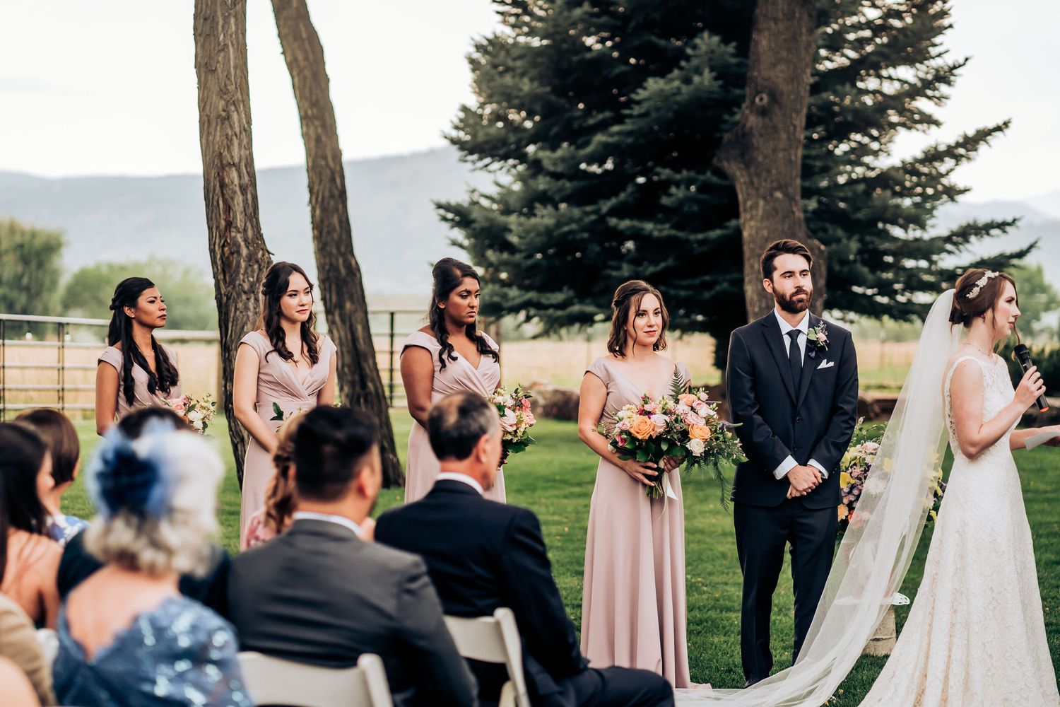 Bridesmaids in blush dresses stand during outdoor mountain wedding ceremony.