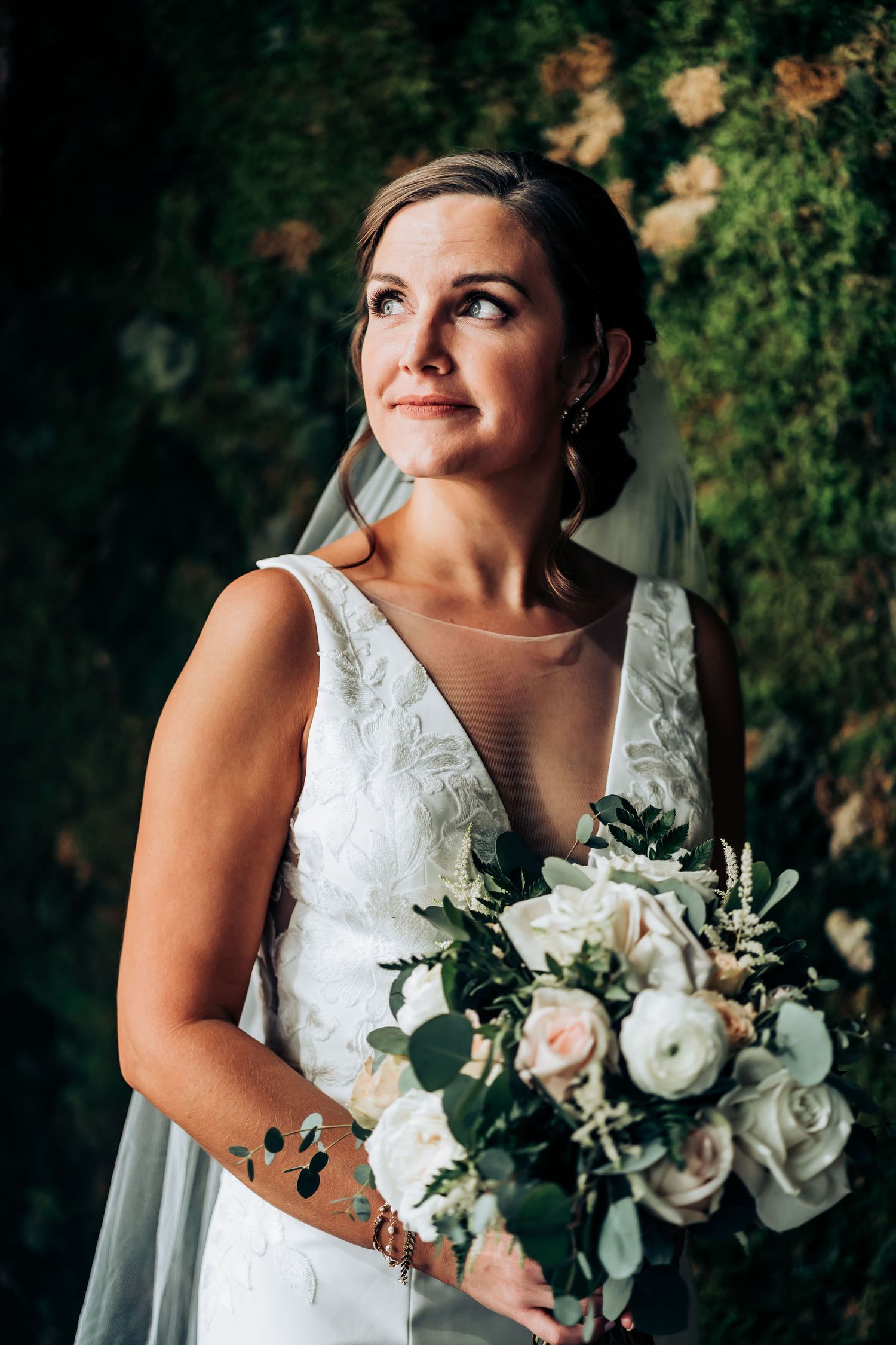 A bride in a classic white wedding gown holds a lush bouquet while looking up with a joyful expression.