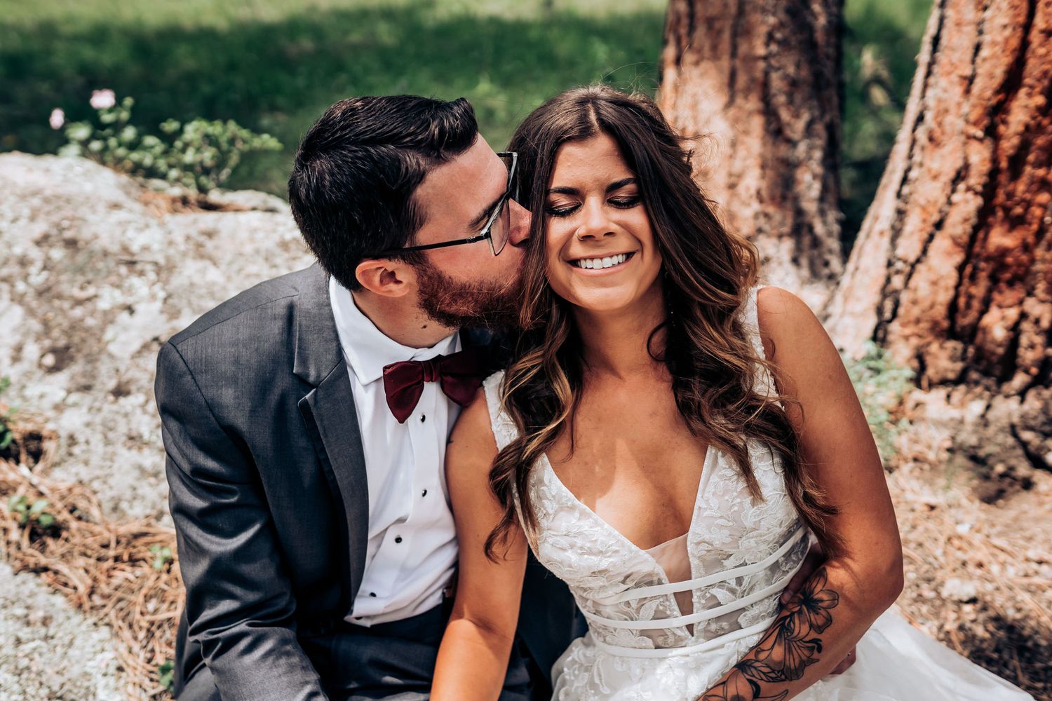 A tender moment captured between newlyweds sitting on a rustic boulder in a forest setting.