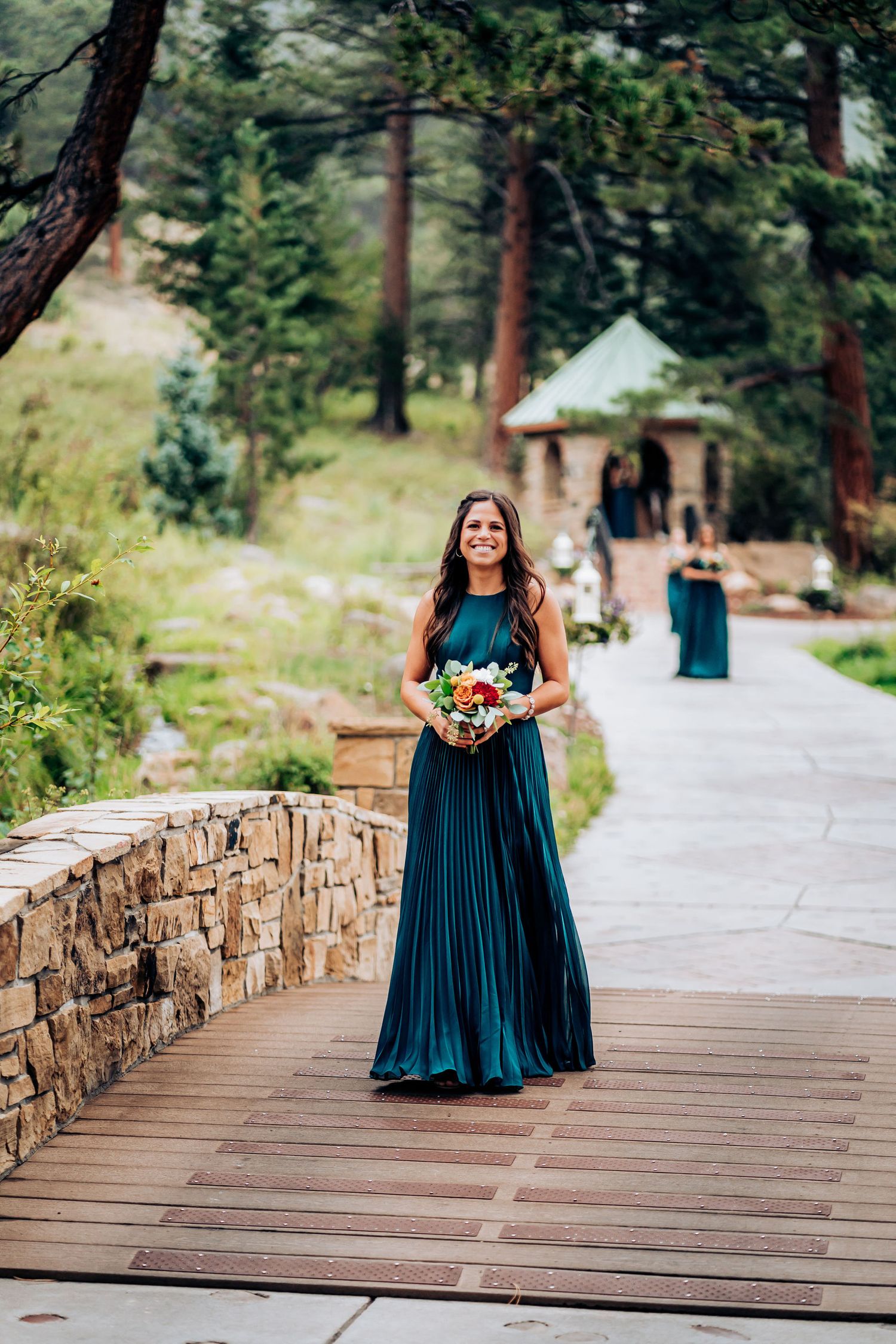Person in flowing teal dress holding colorful bouquet on mountain pathway.