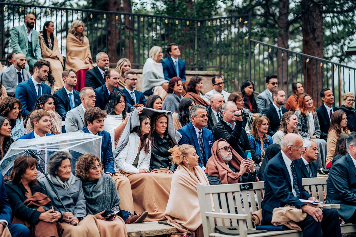 Wedding guests seated outdoors watch ceremony among tall pine trees at mountainside venue.