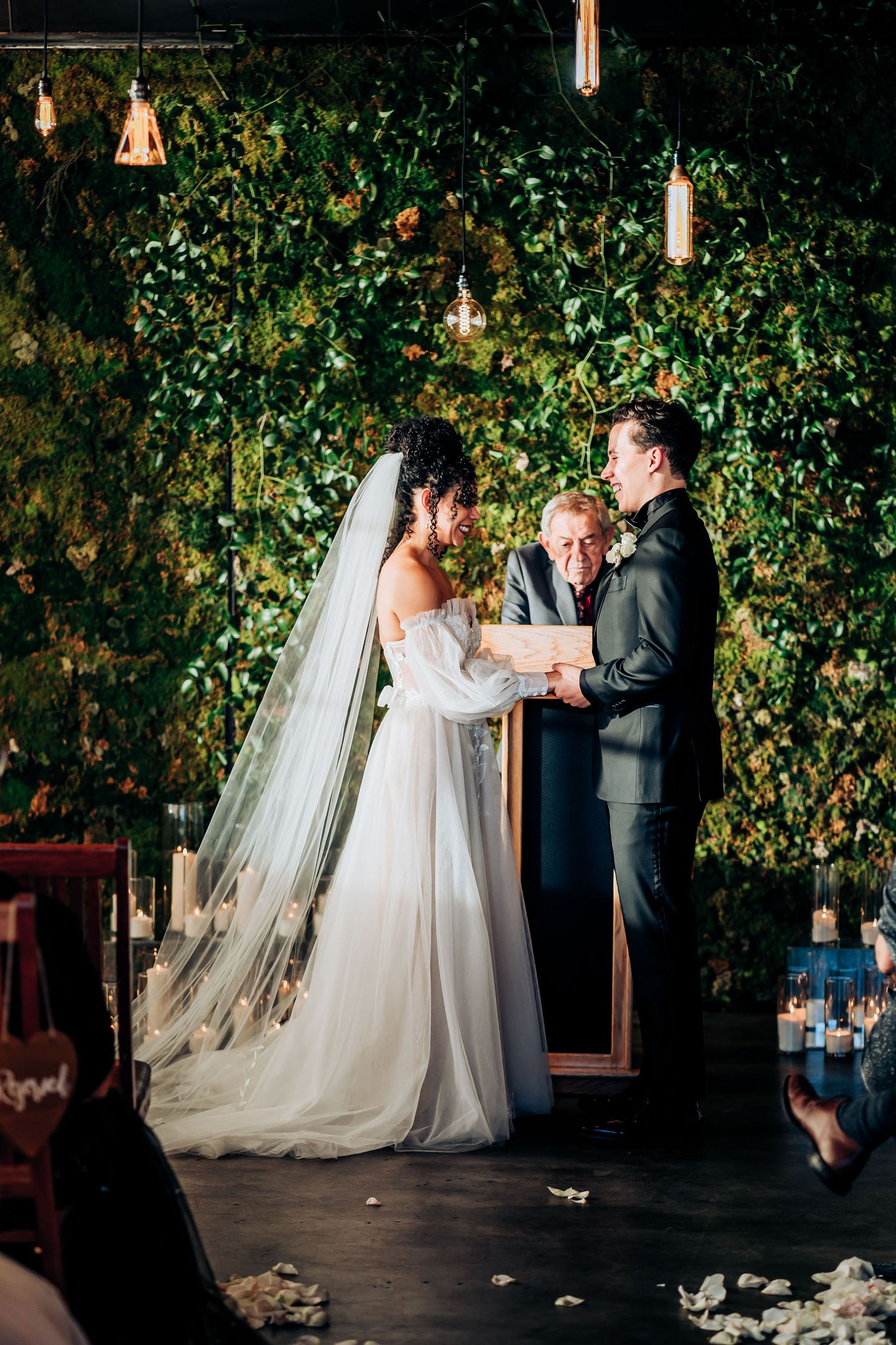Couple exchanging vows during outdoor wedding ceremony with greenery backdrop and hanging lights.
