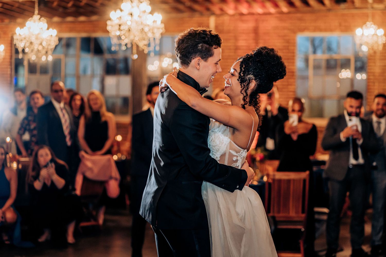 A couple shares their first dance under crystal chandeliers while guests watch in an elegant venue.