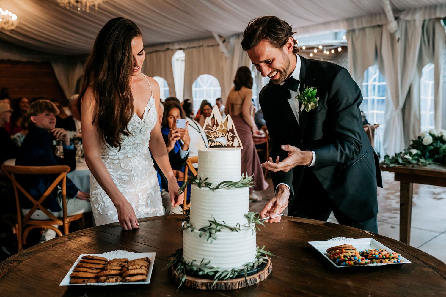 Elegant three-tiered white wedding cake being cut at a reception with greenery decorations.