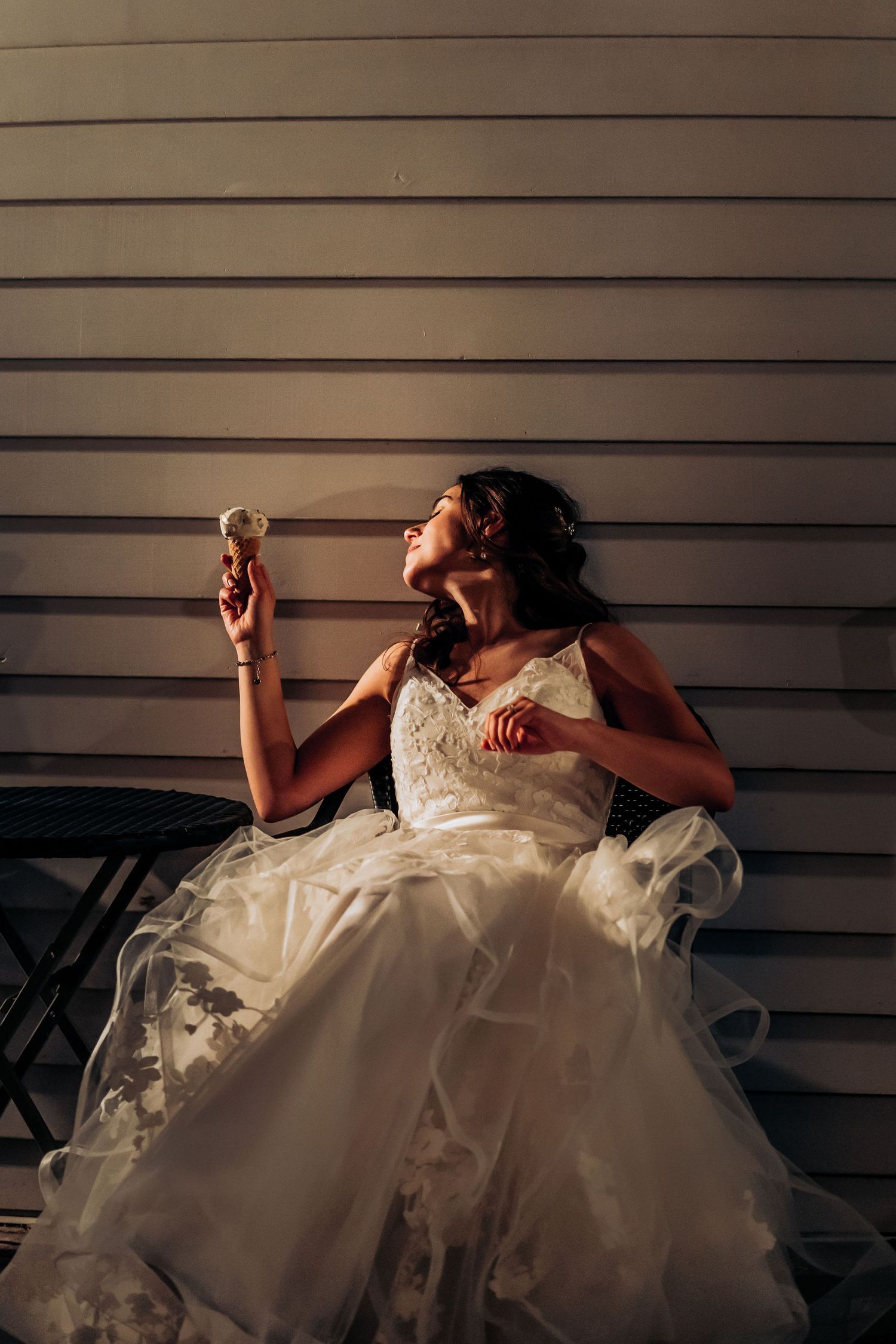 Bride in white dress poses dramatically while enjoying ice cream cone against wall.