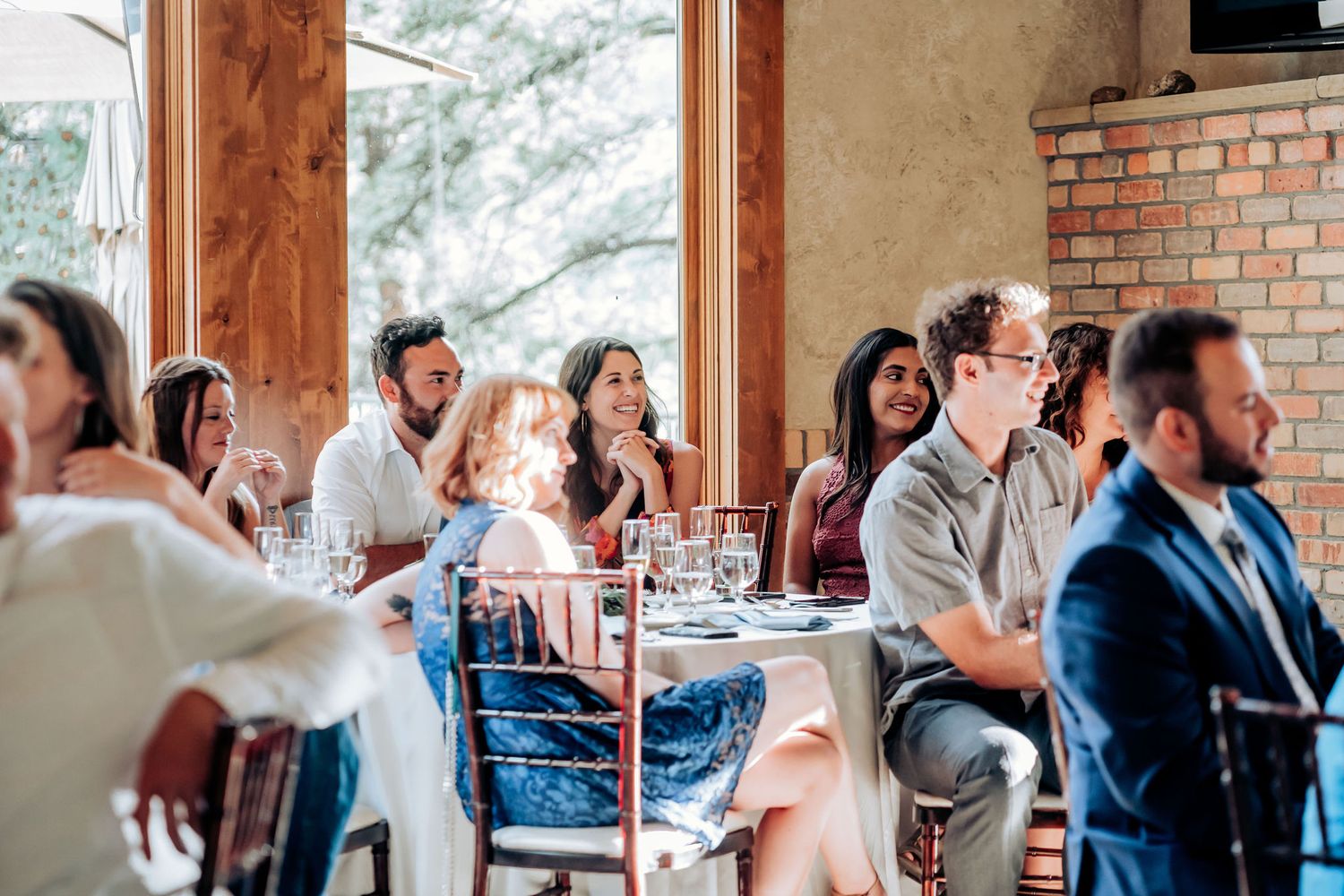 Wedding reception guests gathered at tables in a rustic venue with wooden beams and brick walls.