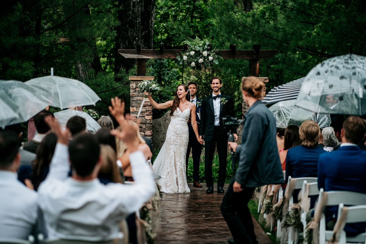 Wedding guests hold umbrellas during an outdoor ceremony on a rainy day.