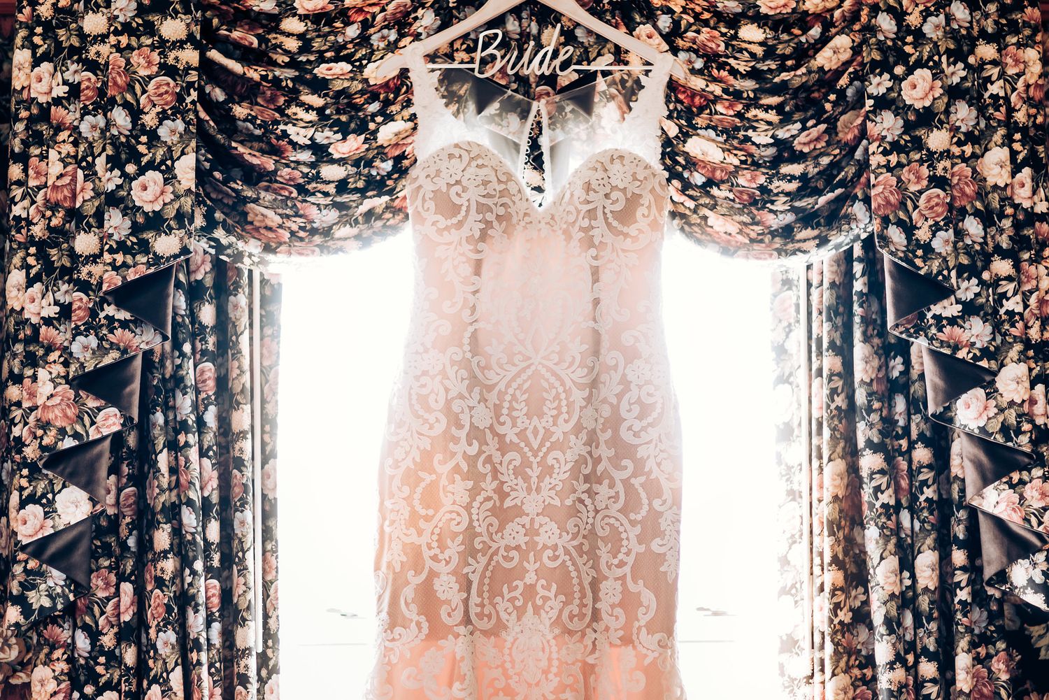A delicate lace wedding dress hanging in front of floral curtains with natural light streaming through.