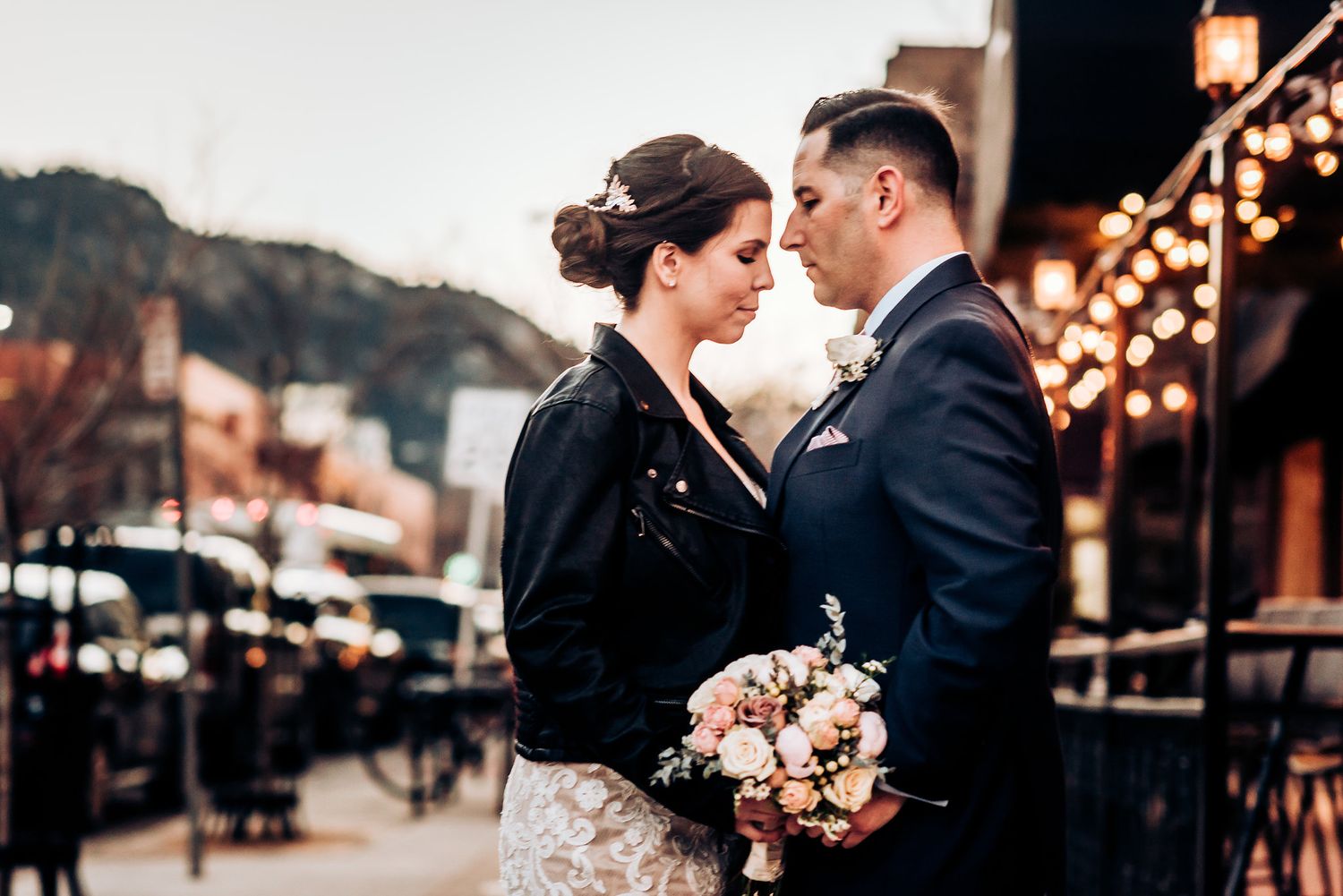 A couple embraces outdoors with string lights and a bouquet of pink flowers.