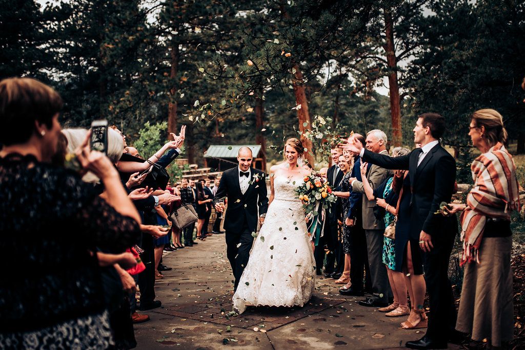 Wedding guests celebrate by showering flower petals on a newlywed couple along a forest path.