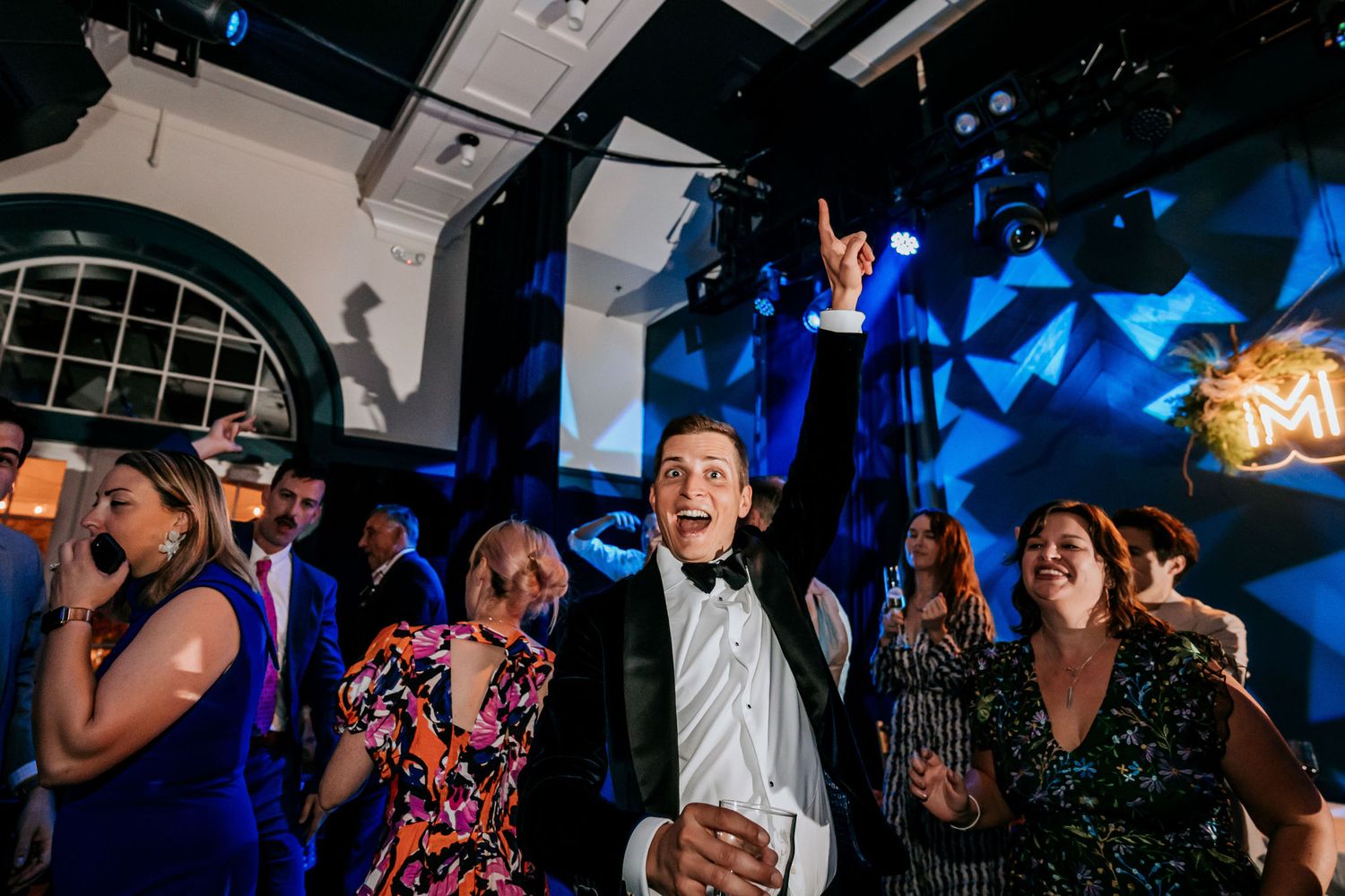 Party guests dance enthusiastically under blue lighting at an indoor celebration venue.