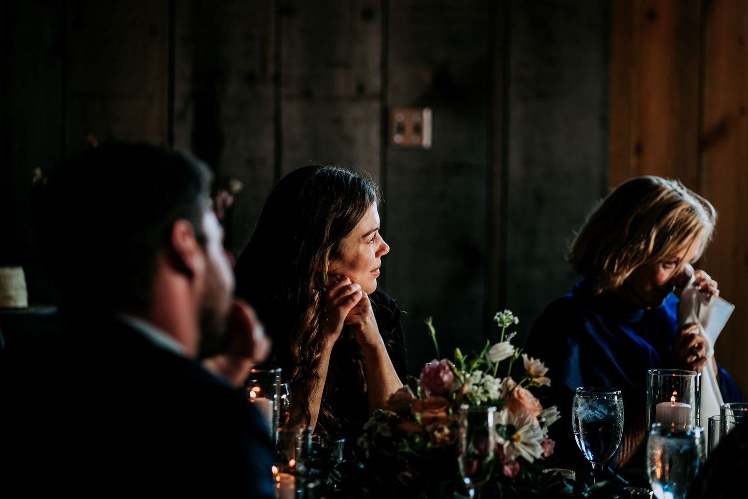 Guests share emotional reactions during dinner speeches in a dimly lit reception venue.