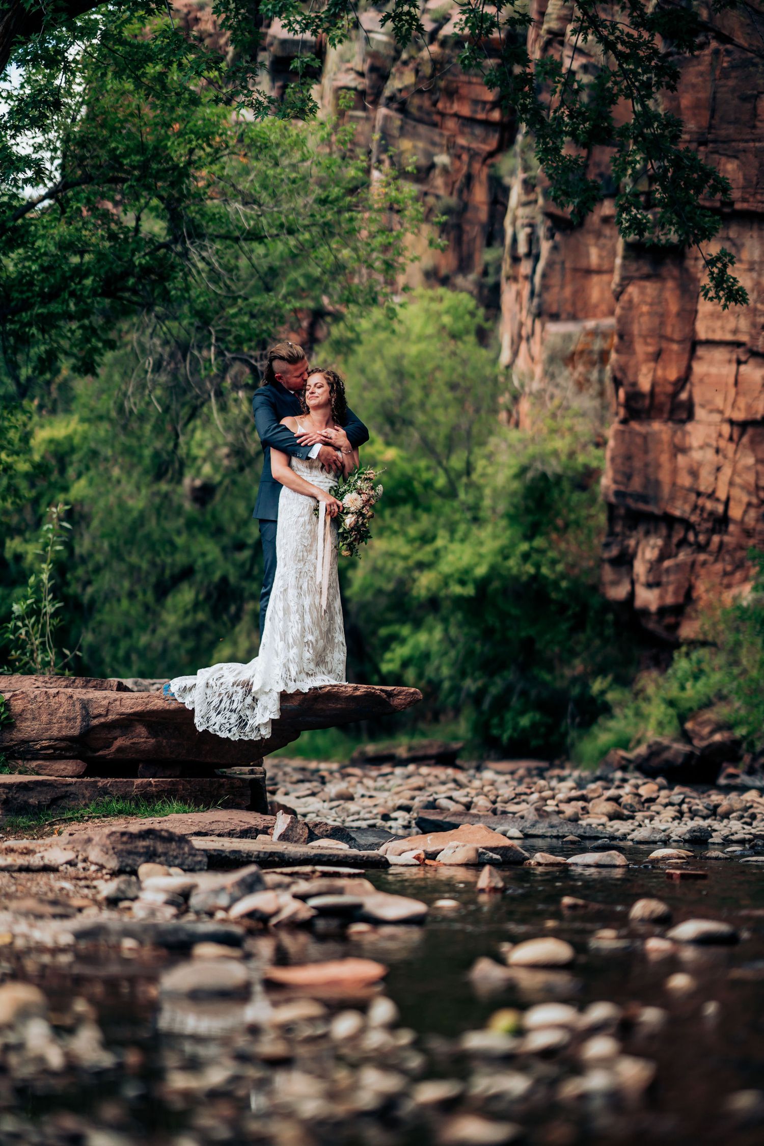 Wedding couple embraces on rocky ledge overlooking river with dramatic cliff backdrop.