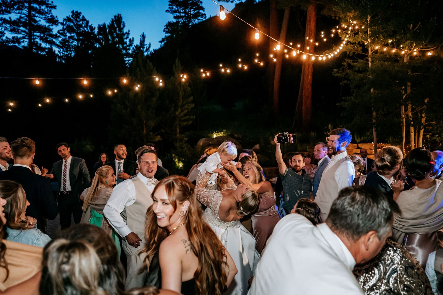 Energetic dance party scene at an outdoor evening wedding reception with string lights.