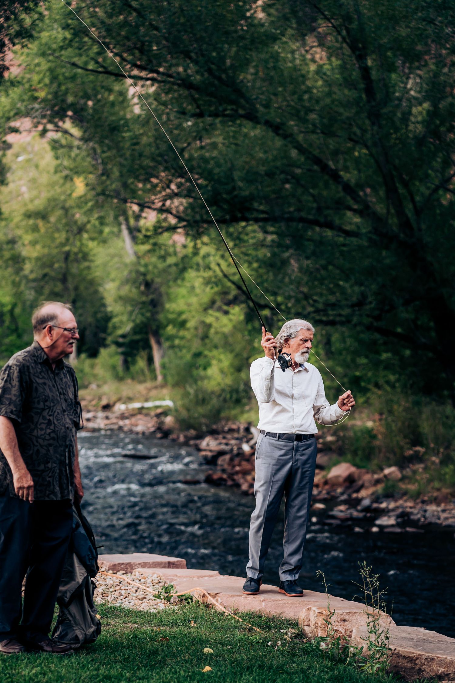 Two people fishing by a mountain stream with lush forest background.