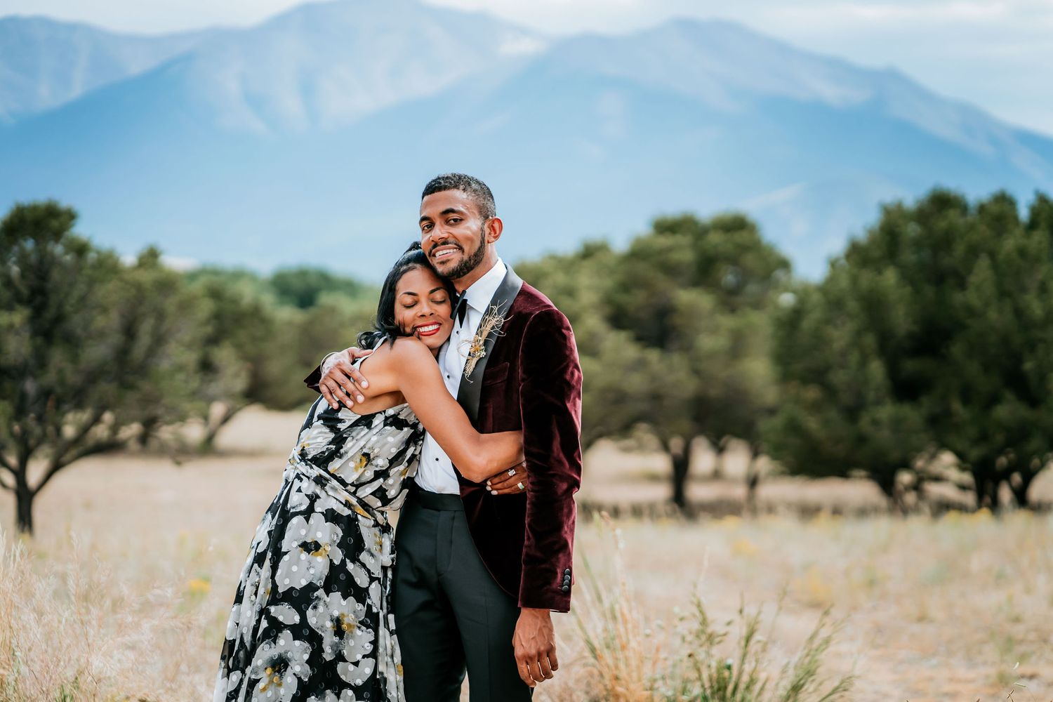 A couple embracing and laughing in a field with mountain views in the background.