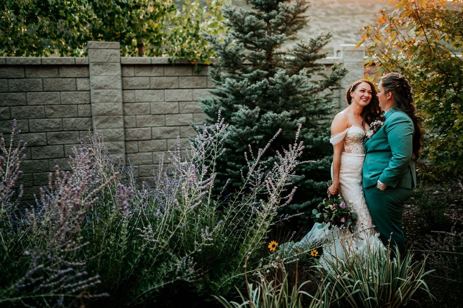 Two brides standing together in a colorful garden with lavender blooms and evergreen trees.