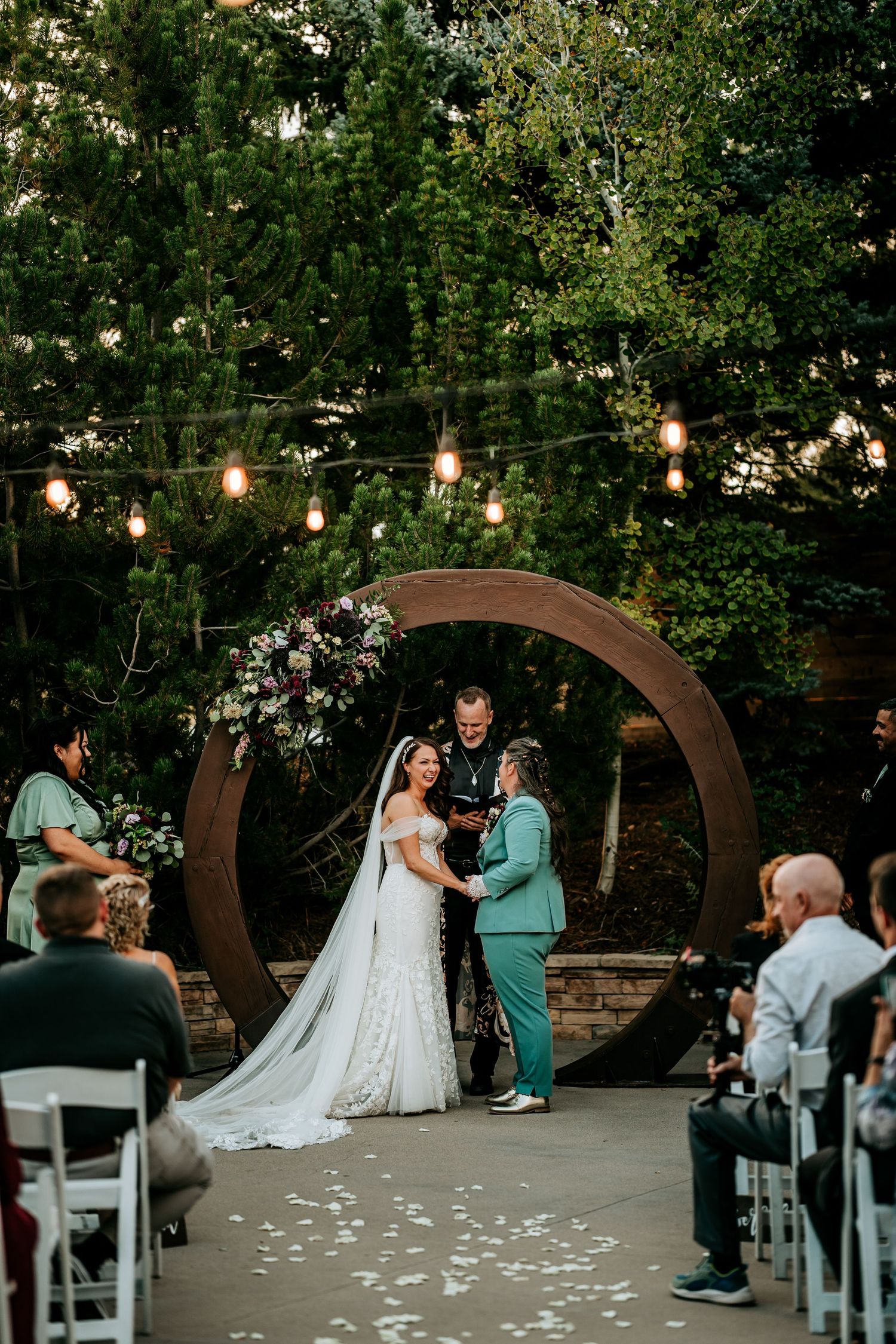 Two brides exchange vows under a circular arch with string lights and floral accents at an outdoor wedding ceremony.