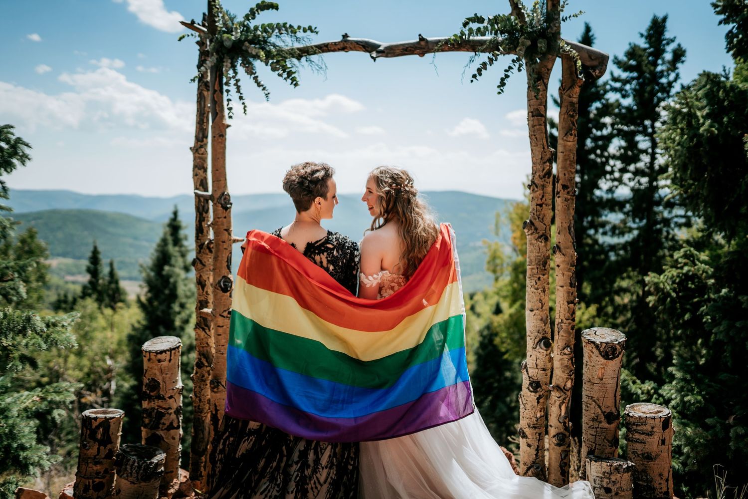A couple holds a rainbow pride flag while standing among tall pine trees with mountains in the background.