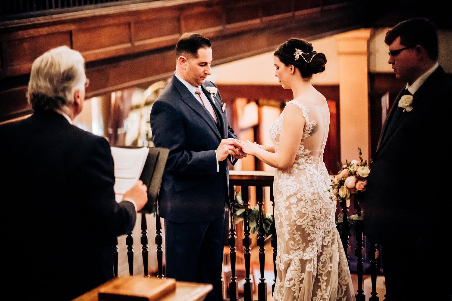 Wedding ceremony captured during ring exchange in an ornate wooden interior setting.
