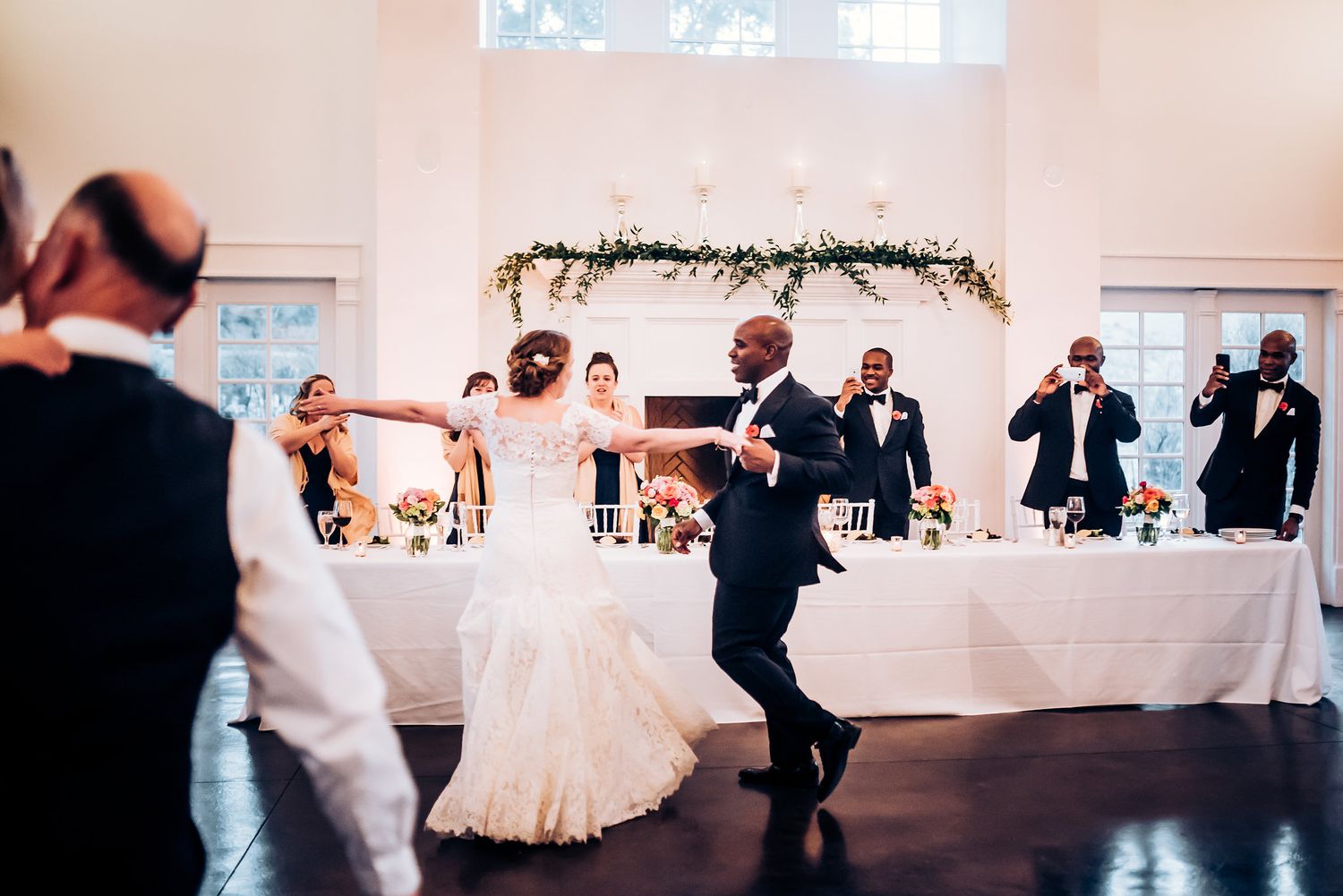 Energetic dance scene at wedding reception with decorative garland and white backdrop.