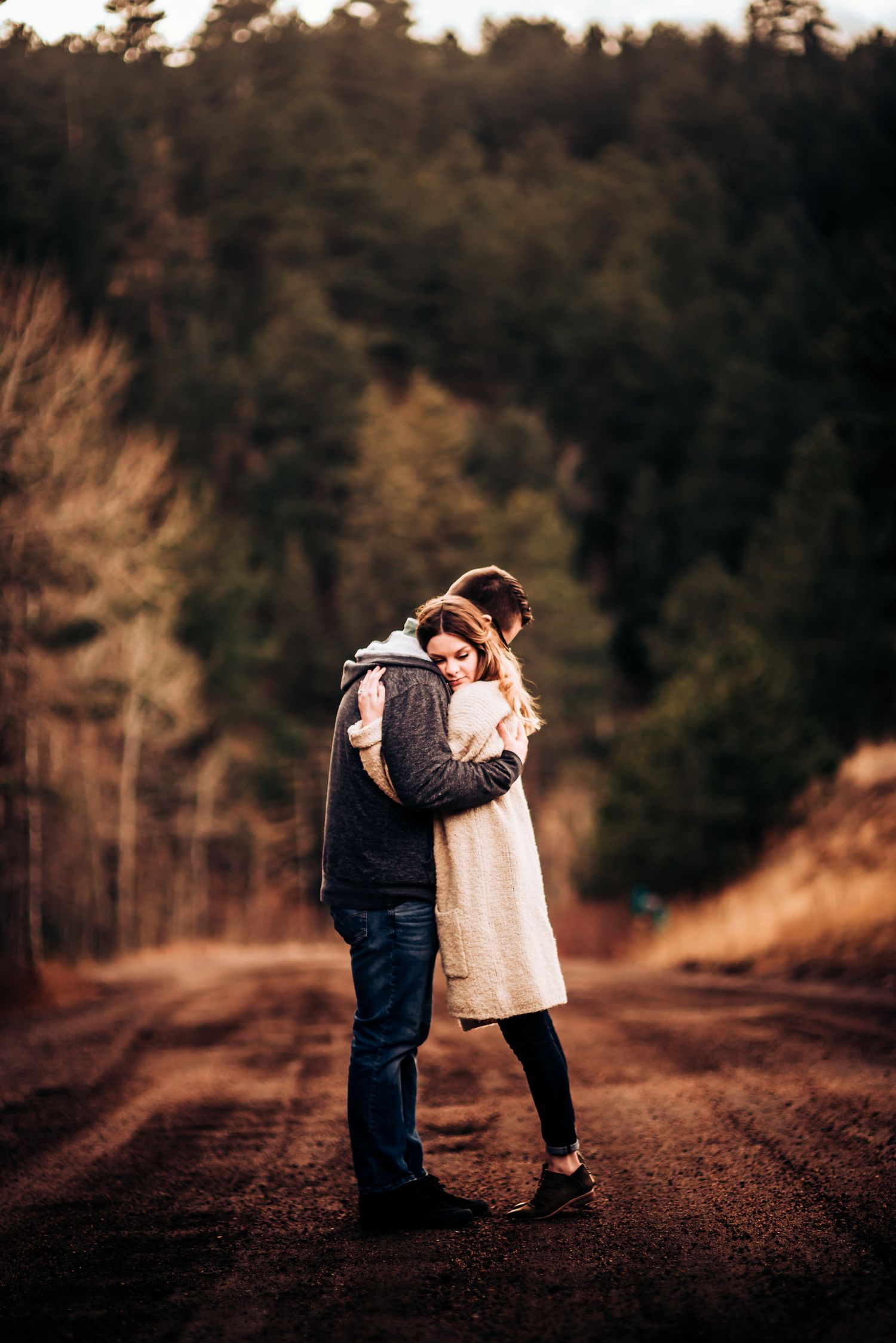 Couple sharing intimate moment while standing on dirt road surrounded by forest.